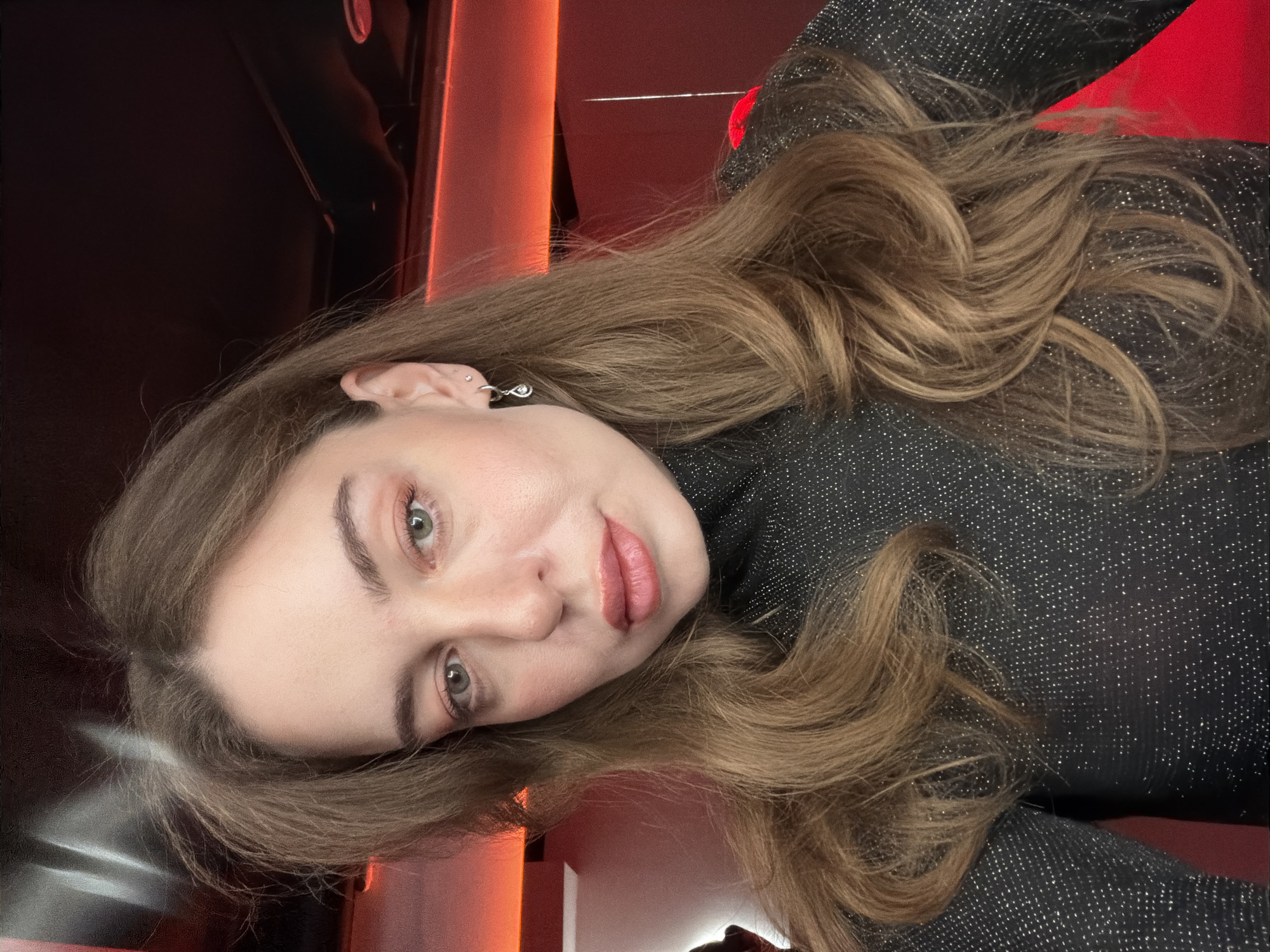A close-up, head-and-shoulders portrait of a light-skinned female individual with long, wavy brown hair and green eyes. She is wearing a black turtleneck top flecked with shimmering particles. Her visible right ear features two earrings: a small stud and a teardrop-shaped dangle earring. Her face exhibits light makeup, including peach-toned eyeshadow and pink lipstick. The individual gazes directly forward, slightly angled towards the camera. The background is dimly lit with a prominent, warm red glow emanating from the right side, illuminating surfaces in a deep red hue, while the upper background and ceiling are dark. This scene is situated in Moscow, Russia.