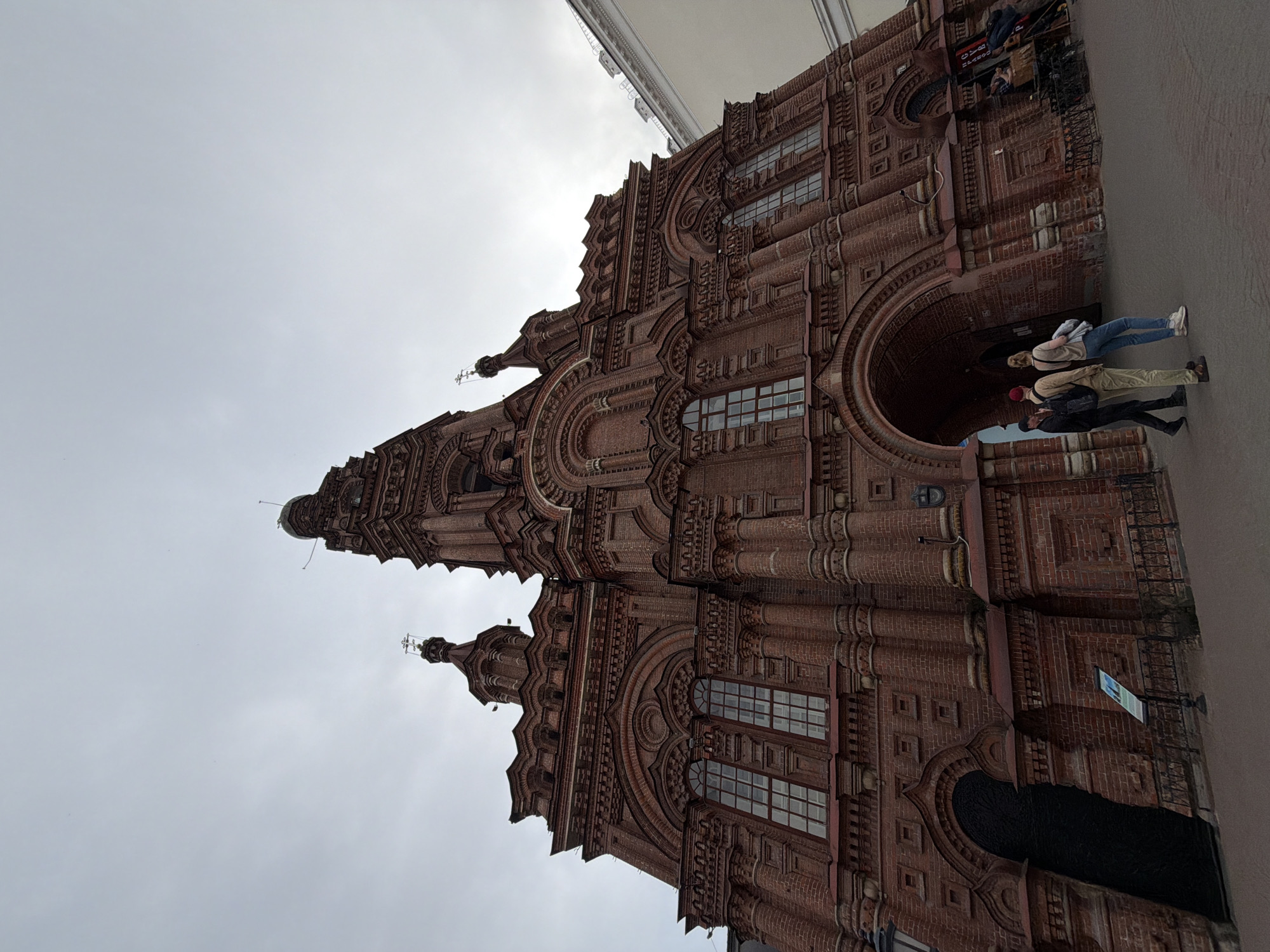 Here is a detailed description of the image:
The image shows the Cathedral of the Annunciation, located in Kazan, Russia. The cathedral is a red brick building with several arched windows and doorways. The style is Russian Revival. There are several people standing near an arched doorway. The sky is overcast.