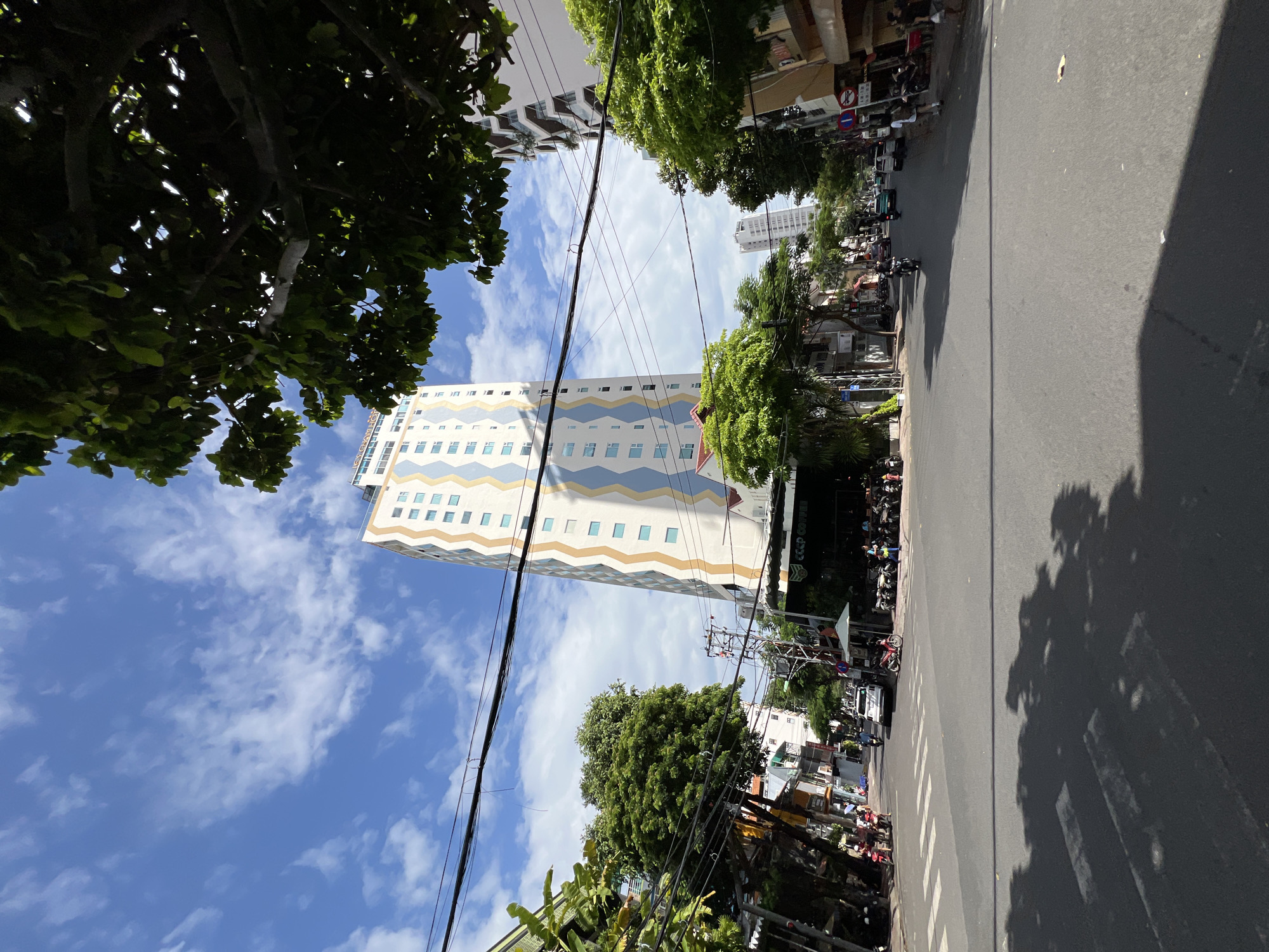 This is a street-level, eye-level, wide-angle shot taken on a sunny day in Nha Trang, Vietnam. The scene captures a typical urban streetscape with a prominent tall building with a distinctive zigzag pattern of blue and gold on its facade, standing out against a bright blue sky with scattered white clouds. The building appears to be a hotel or apartment complex.
In the foreground, a paved road with white pedestrian crossing lines dominates the lower half of the image. Shadows of trees are cast across the road, indicating the presence of lush greenery. On the left side of the street, there are other buildings, some with trees and balconies, and a narrow sidewalk. On the right side, more buildings line the street, including a cafe with outdoor seating, visible by a sign that reads "CCCP COFFEE".
There are several people and vehicles on the street. On the left sidewalk, a few individuals are standing or walking, and motorcycles are parked. The road itself has a white car driving away from the camera, and several motorcycles are also present, some in motion and some parked. A few people are seen walking on the right sidewalk. Overhead, a network of electrical wires crisscrosses the sky, a common sight in Vietnamese cities. The overall impression is a vibrant, bustling urban environment.