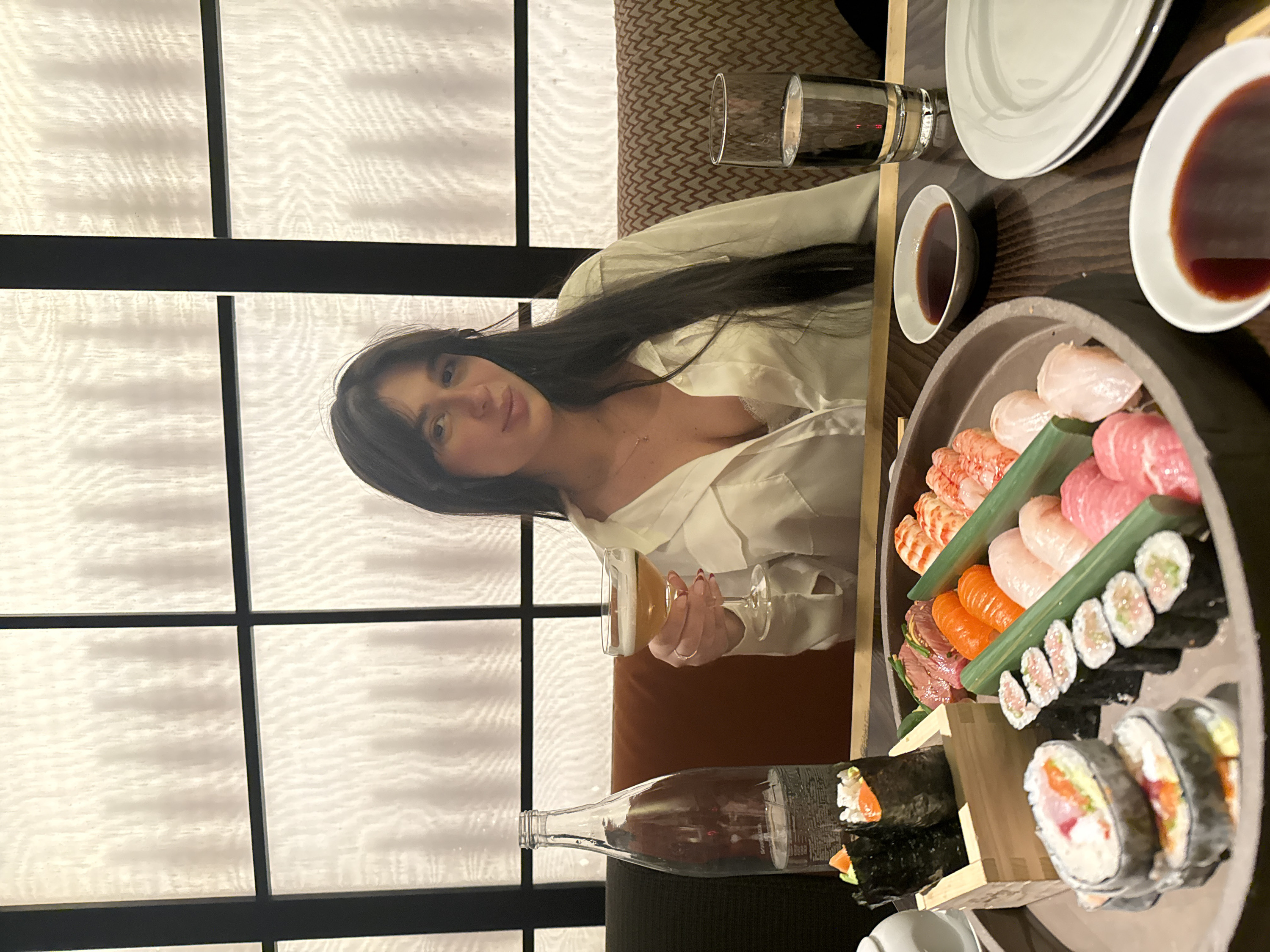 The image captures a dining scene in what appears to be a Japanese restaurant. A young woman with long, dark hair and bangs is seated at a dark wooden table, looking towards the camera with a slight, pleasant smile. She is wearing a light-colored, possibly white, collared shirt, and a delicate necklace is visible around her neck. Her right hand, adorned with red nail polish, holds a stemmed glass containing a light-colored beverage, likely beer or white wine.
In the foreground and to her right, a large, dark-colored platter is filled with an assortment of sushi and sashimi. Various pieces of nigiri are arranged on the platter, including salmon (orange), tuna (pink), white fish, and what appears to be shrimp. Slices of a reddish, thinly cut meat, possibly seared beef, are also part of the selection. Several types of maki rolls, including those with rice on the outside (uramaki) and traditional nori-wrapped rolls, are neatly arranged, some sliced and others whole. Green leaves, possibly bamboo or shiso, are used as garnishes and dividers on the platter.
On the table around the sushi platter, there are two small white bowls filled with a dark sauce, presumably soy sauce, and a larger empty white plate. A clear glass of water and a tall, clear glass bottle (possibly containing water or sake) are also visible on the dark table surface.
The background features a light-colored partition or screen with a grid pattern, consisting of dark horizontal and vertical bars separating translucent, subtly textured panels. This design element reinforces the aesthetic of a Japanese dining establishment. The lighting in the scene appears to be soft and ambient, suggesting an evening meal. There is no visible text in the image. The overall impression is one of a relaxed and enjoyable dining experience.