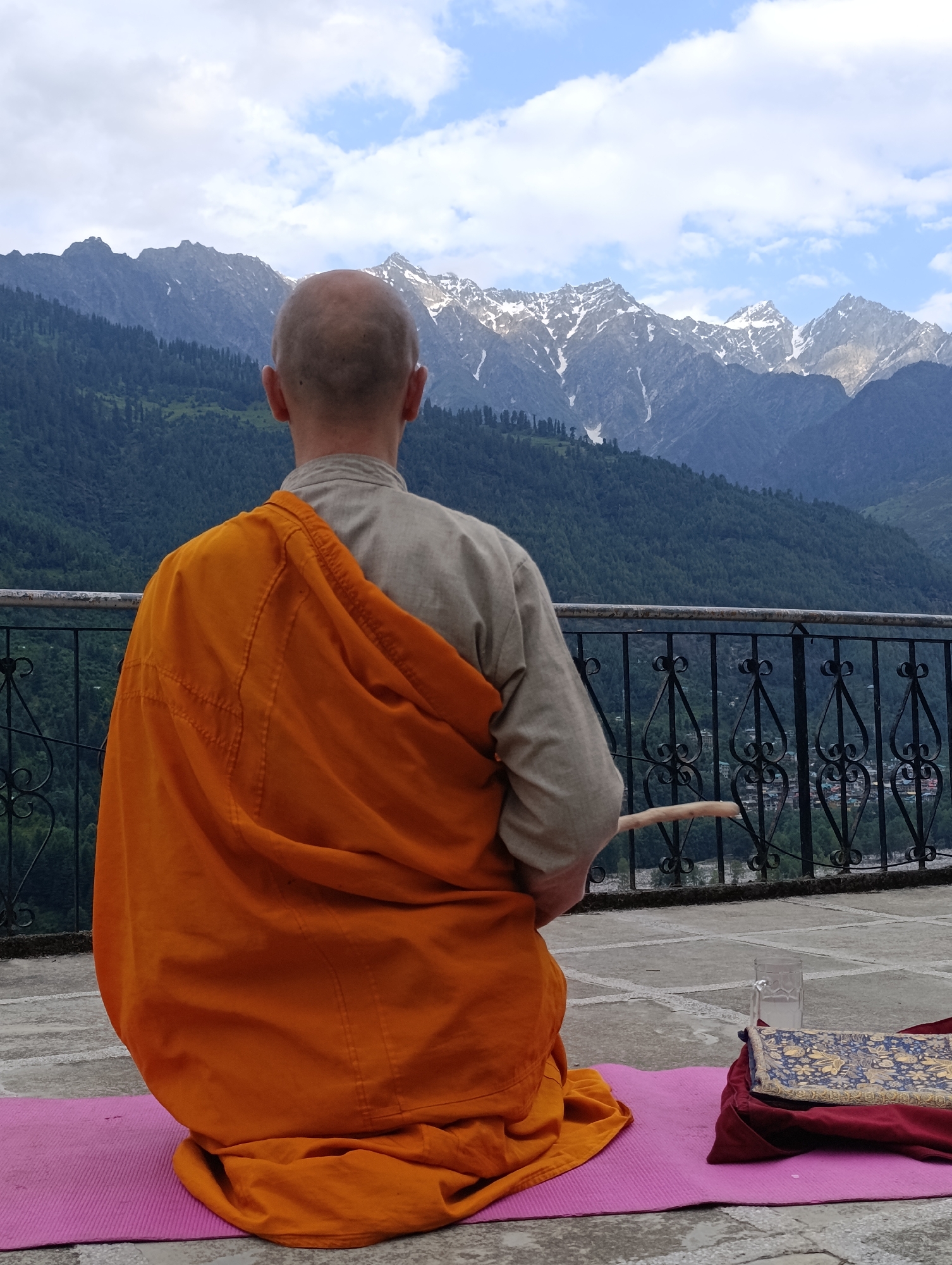 An overhead shot shows a bald man from the back wearing an orange robe and a light-colored shirt, sitting cross-legged on a pink yoga mat. He is facing a mountain range, with snow-capped peaks in the distance. A wooden stick is held in his right hand. To his right is a glass of water and a blue and gold patterned book. The man is sitting on a paved area with a black metal railing in front of him, separating him from a lush green valley below. The sky above is cloudy.
