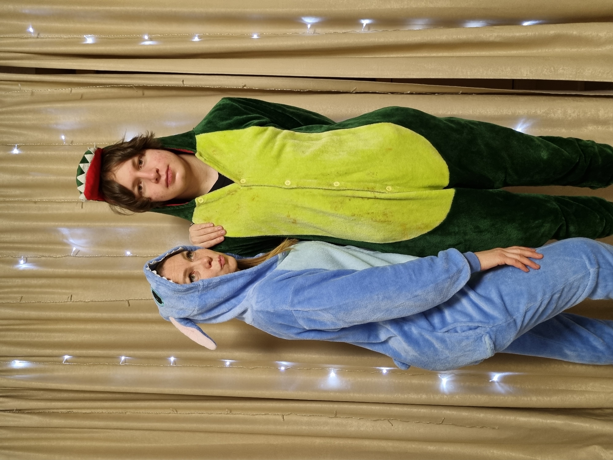 Two individuals, a young man and a young woman, are depicted lying side-by-side in a cozy indoor setting in Chaikovsky, Russia. The background consists of light brown or beige curtains, draped with horizontal folds, and adorned with several strands of white string lights that emit a soft glow.
The young man, on the left, is wearing a dark green and lime green kigurumi (animal-themed onesie) that resembles a dinosaur or dragon, complete with a hooded head featuring red, jagged spikes. He has medium brown, wavy hair and looks directly at the camera with a neutral expression.
The young woman, on the right, is dressed in a light blue and blue kigurumi, designed after the character Stitch, identifiable by its prominent ears on the hood. Her hair is light brown, and she also looks towards the camera with a neutral gaze.
Both subjects appear relaxed and comfortable in their full-body costumes, suggesting a playful or casual event, possibly an evening gathering or simply a comfortable night in. No specific activity beyond posing is evident. The string lights contribute to a warm, ambient atmosphere. There is no visible text in the image.