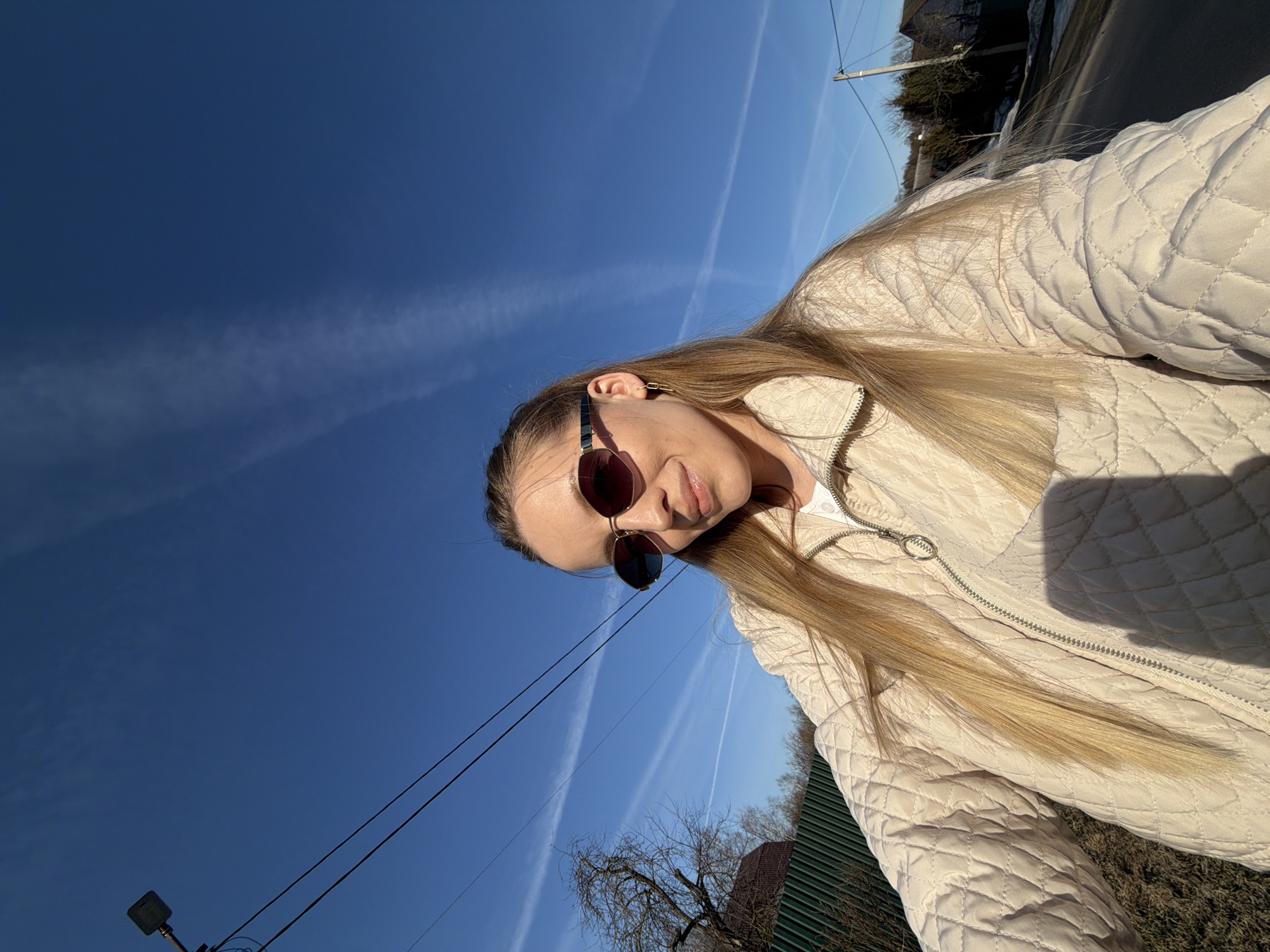 A woman with long blonde hair and sunglasses stands outdoors in Reshetnikovo, Russia. She is wearing a light-colored quilted jacket. The sky is blue with contrails. Power lines are visible in the foreground. A house with a green fence and a bare tree are in the background.