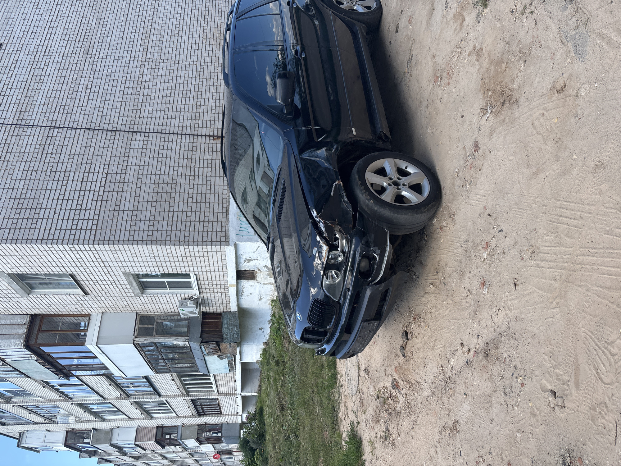 A black BMW X5 SUV is parked on a dirt and gravel lot next to a multi-story brick apartment building. The front passenger side of the SUV is visibly damaged, with the fender, headlight, and bumper appear to be deformed and broken. The tires on the front passenger side are deflated. The apartment building has balconies with glass enclosures and some appear to be in disrepair. The ground is uneven and covered in dirt, small stones, and some patches of dry grass. The sky is overcast. This scene is located in Kstovo, Russia.