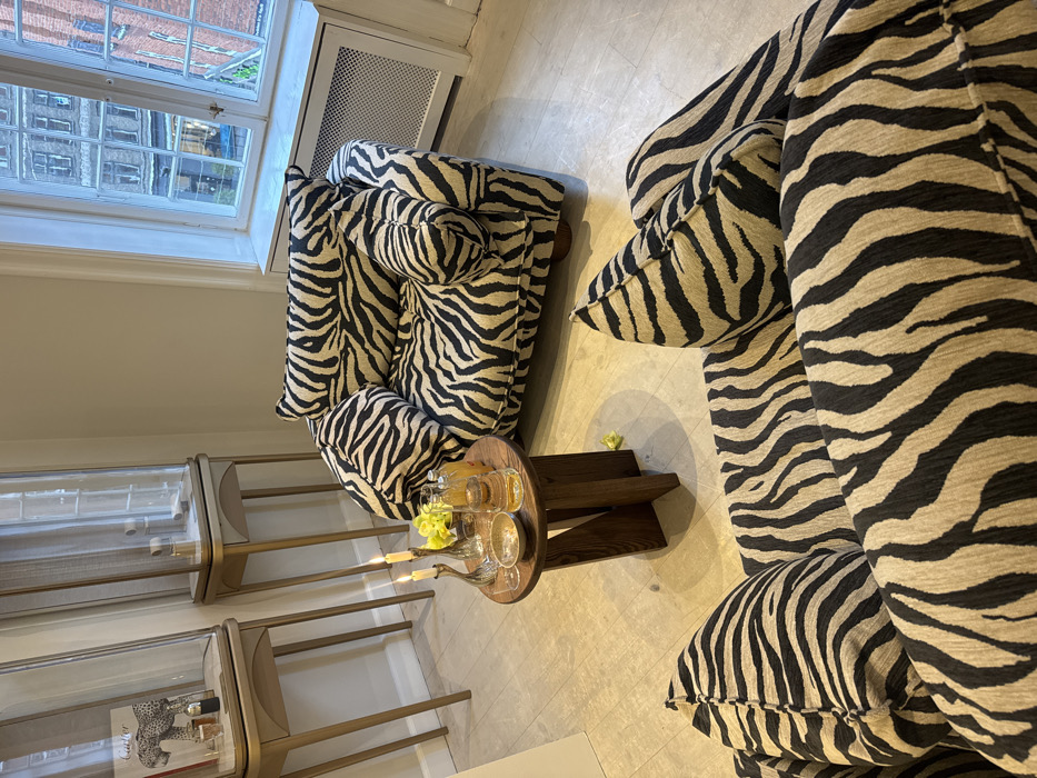 The image displays an interior room, likely a commercial lounge or showroom, situated in a city (Unknown, Unknown). The space features light-colored walls and flooring. Two pieces of furniture, an armchair positioned facing a window and a section of a sofa or second armchair in the foreground, are upholstered in a beige and black zebra-print fabric. A circular dark wood side table stands between the seating, holding two lit candles in silver-colored holders, a clear glass bottle with orange liquid, two drinking glasses (one containing an amber liquid), and a small textured bowl. Scattered yellow decorative items are visible on the table and floor. Along the left wall, two tall, narrow display cases with glass tops and light-colored bases showcase items; the left case contains a book with a leopard cover and small dark objects, while the right contains smaller light-colored objects on stands. A large window on the right reveals an exterior urban view of multi-story brick buildings with multiple windows. A white radiator cover is present below the window. No individuals are depicted.