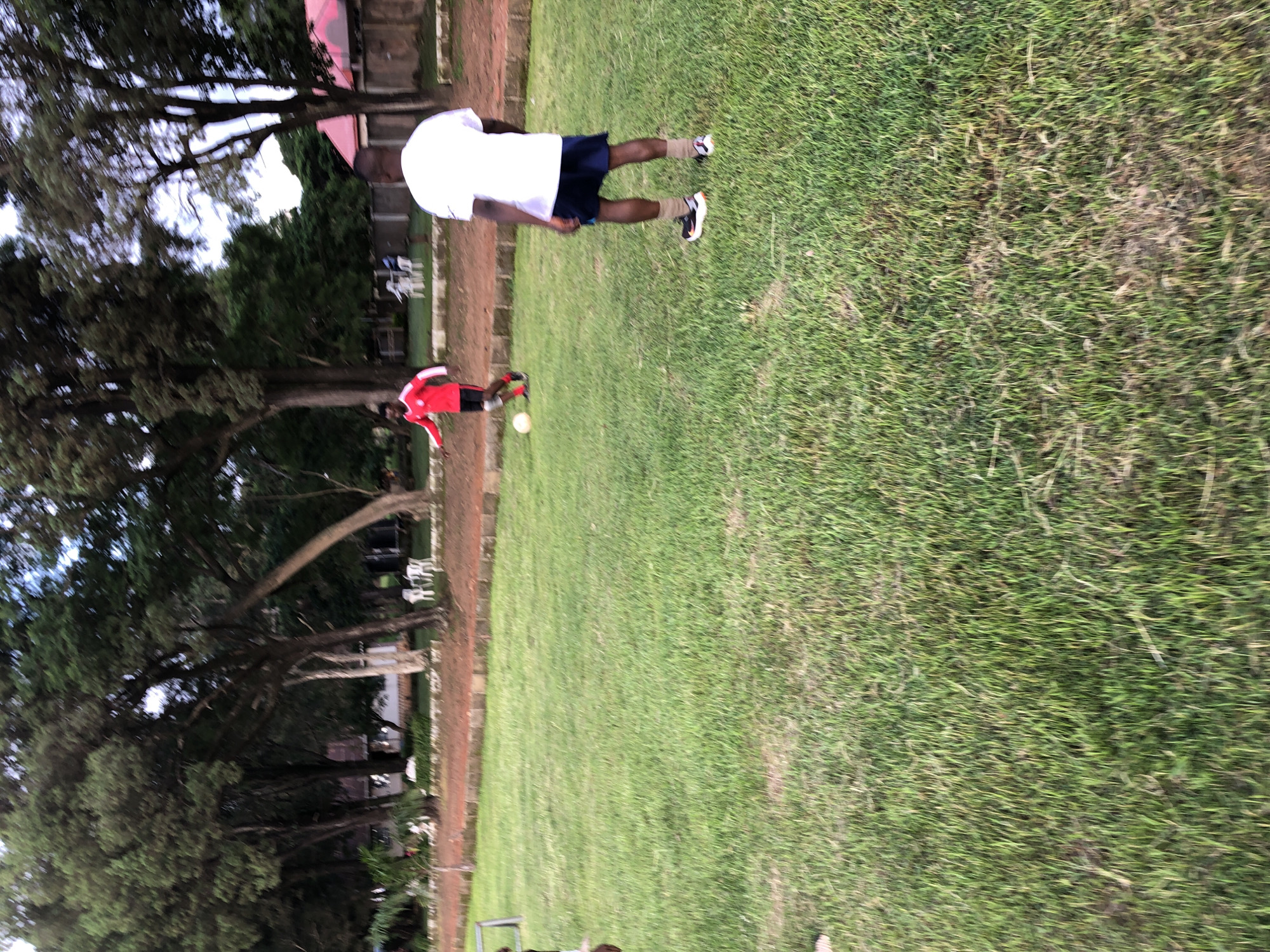 The image depicts two individuals on a grassy field, possibly a sports field, given the presence of a soccer ball near one of the subjects. One person, wearing a white t-shirt and dark shorts, is standing on the grass. The other individual, in a red and black athletic outfit, is positioned lower in the frame and appears to be interacting with the soccer ball. The setting includes trees and a building with a red roof in the background, suggesting an outdoor environment. The lighting suggests it is daytime. There is no visible text in the image. The overall scene appears to be a casual sports activity.