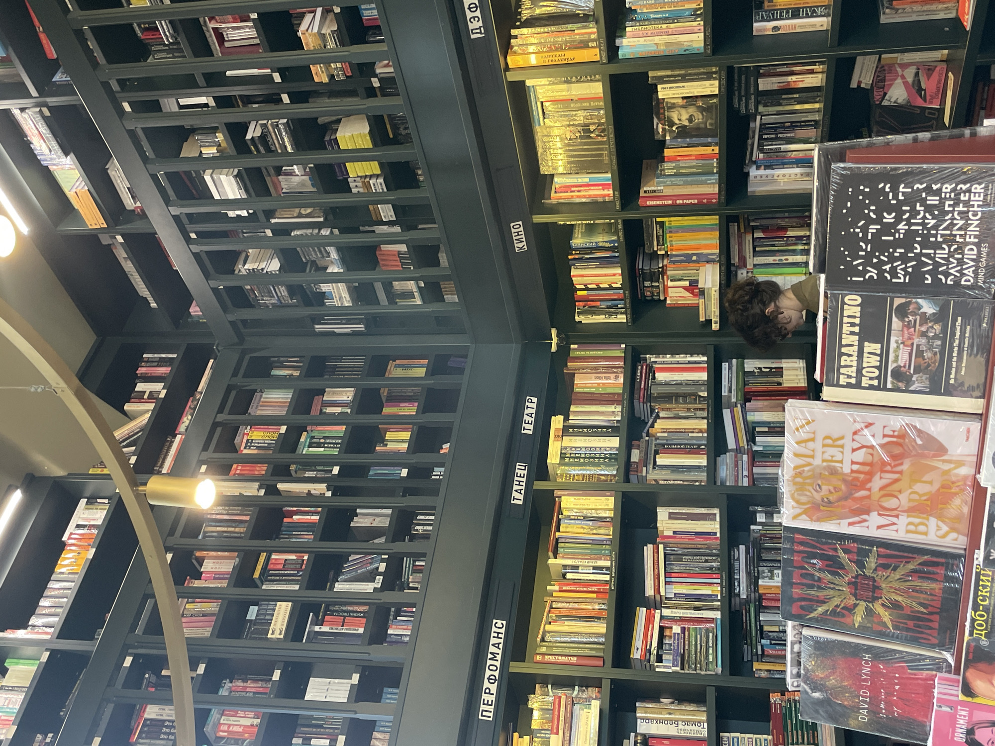 The image shows the interior of a bookstore in Saint Petersburg, Russia. The store features tall bookshelves reaching towards the ceiling, filled with books. The shelves are labeled with categories such as "Performans" (Performance), "Tanets" (Dance), "Teatr" (Theater), "Kino" (Cinema), and "Def" (Def). The books are arranged neatly on the shelves. A person with curly hair is partially visible between the books. The foreground includes several books prominently displayed, including biographies of Marilyn Monroe and books related to directors David Fincher and David Lynch.