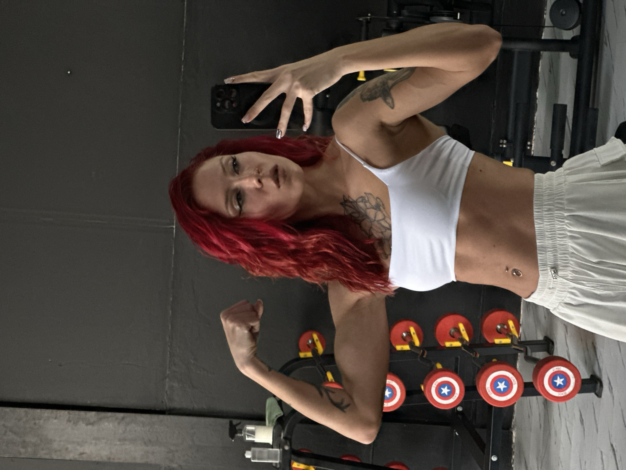 The photograph captures a woman with red hair, dressed in a white crop top and white pants. She's holding a black smartphone, seemingly taking a selfie. The background appears to be a gym setting, featuring workout equipment including a rack holding weights adorned with a Captain America shield design. The location is North Kuta, Indonesia.