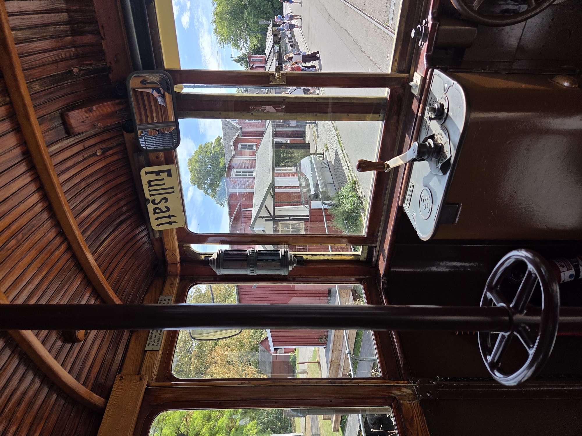 The image is taken from inside a vintage tram, looking out through the driver's cabin window. The interior is made of dark wood, with a curved wooden ceiling and a central vertical pole. A steering wheel and a control panel with levers are visible in the foreground. Outside the tram, a cobblestone street runs alongside tram tracks. Red buildings with white windows line the street, and several people are walking along the sidewalk. Trees and a blue sky with clouds are visible in the background. A sign above the window reads "Fullsatt" in yellow lettering. Based on the signage and architecture, the location is likely in Malmö, Sweden.