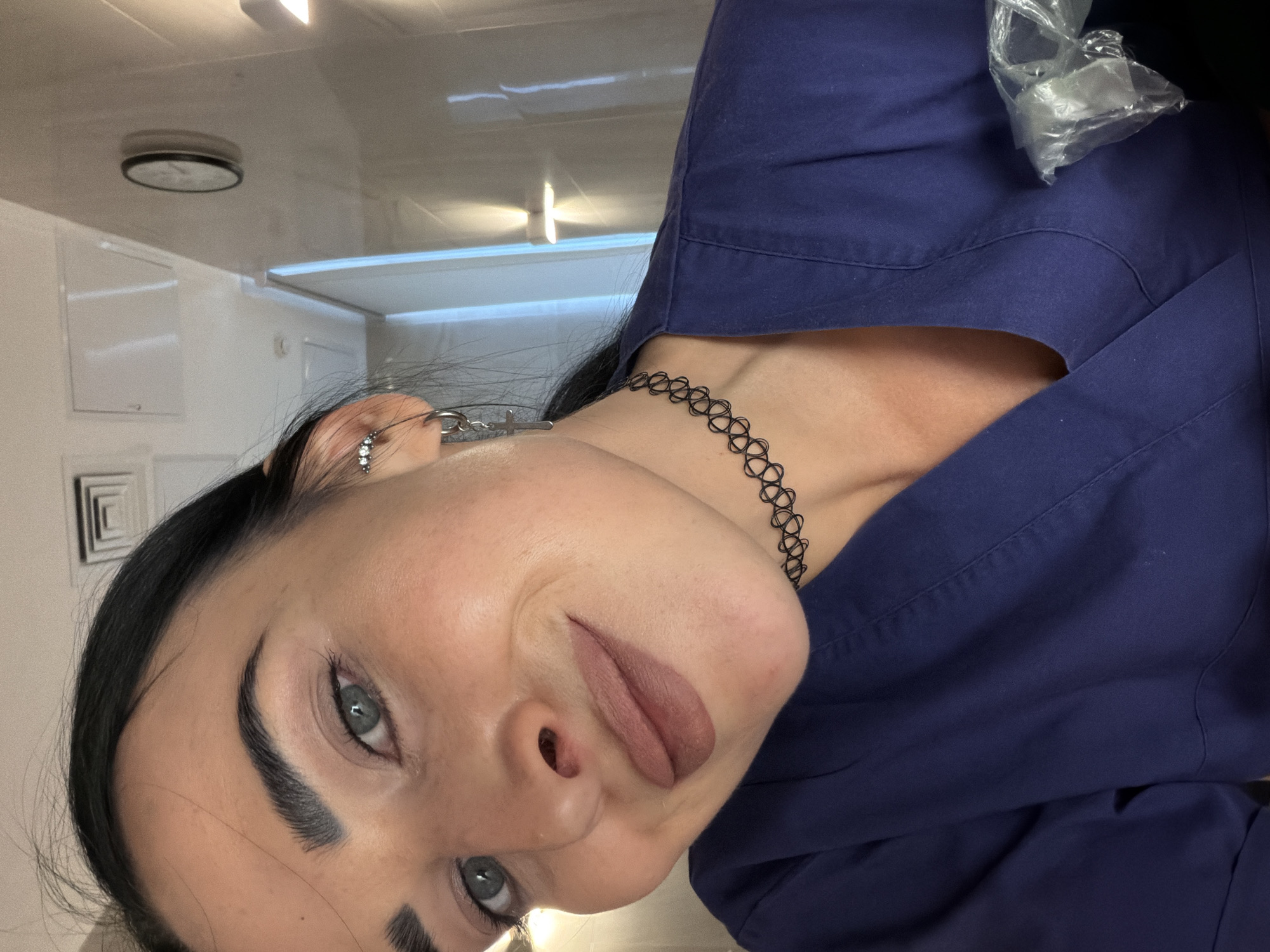 This is a close-up, tilted selfie of a young woman with dark hair, light blue-green eyes, and distinct, well-groomed eyebrows. She looks directly at the camera with a subtle, composed expression, sporting a matte, muted pinkish-brown lipstick.
She is wearing a dark blue, possibly V-neck, garment, which appears to be a uniform or scrubs. Around her neck, she has a black stretchy choker necklace. On her visible left ear, she has several earrings, including a dangling one shaped like a small cross or dagger, and a stud with multiple small stones on her earlobe. A clear plastic bag is visible resting on her right shoulder area, near the top of her blue attire.
The setting is indoors, featuring plain white walls and a white ceiling. A circular ceiling light fixture is visible in the background, along with other recessed lighting and a rectangular panel or vent on the wall. The environment suggests a clean, possibly professional or institutional space like a clinic, office, or restroom. There is no visible text in the image. This image was taken in Saint Petersburg, Russia.