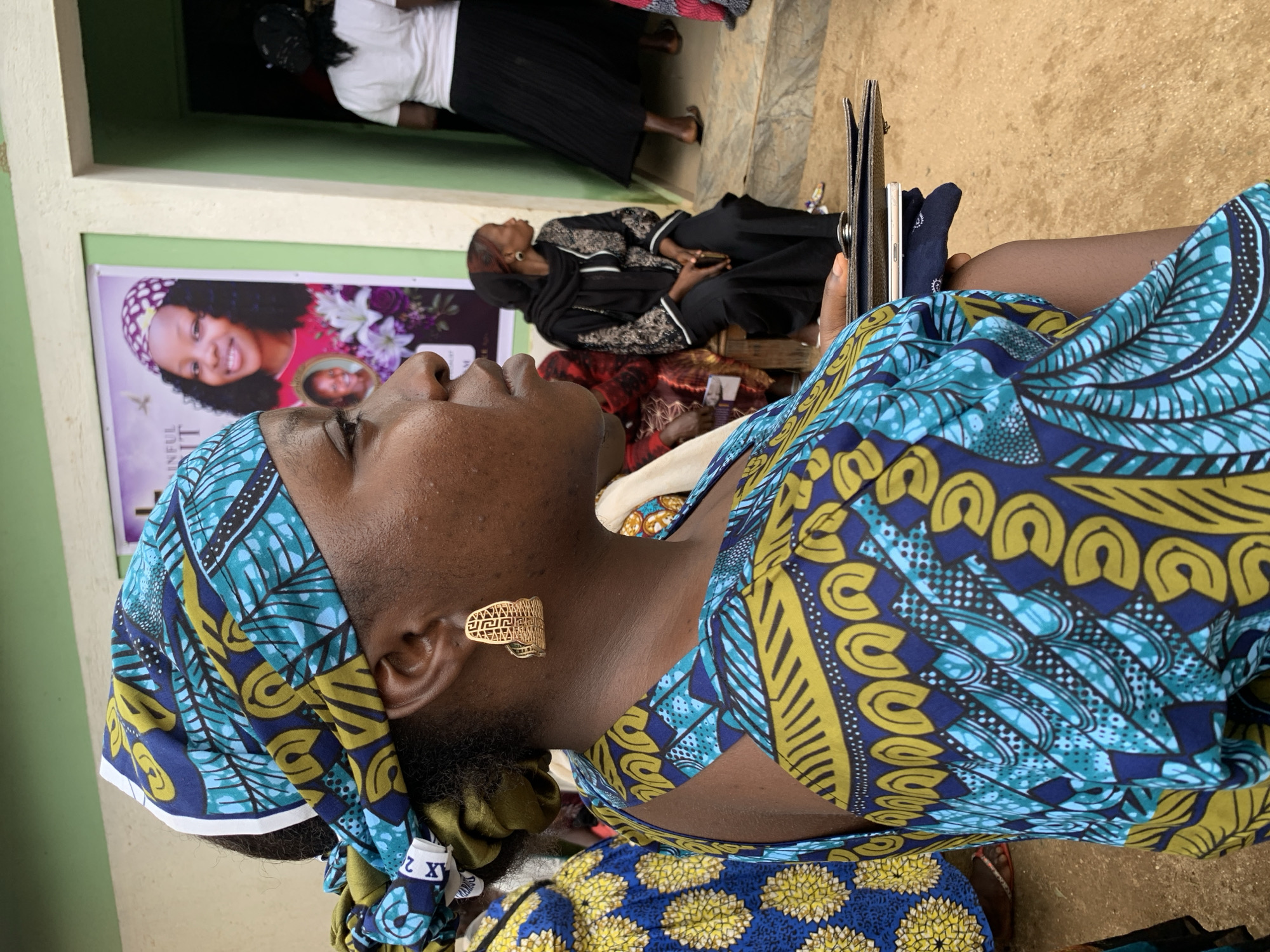 In Miango, Nigeria, a woman is captured in a medium shot, her profile facing right. She wears a blue and yellow patterned headwrap and a matching patterned dress with cut-out shoulders. A large, ornate gold earring adorns her right ear. She holds a wallet and a mobile phone in her right hand, resting them on her left arm, which is positioned across her body.
In the background, to the left, a poster displays a portrait of a smiling young woman wearing a purple headscarf. To the right, a doorway reveals the back of a woman in a white top and a long black skirt, and further back, another woman in a black headscarf and dress sits on a wooden bench. The ground is a dusty, light brown earth. The building behind the subjects is painted a light cream color with green trim around the doorway. The overall scene suggests an outdoor gathering or event.