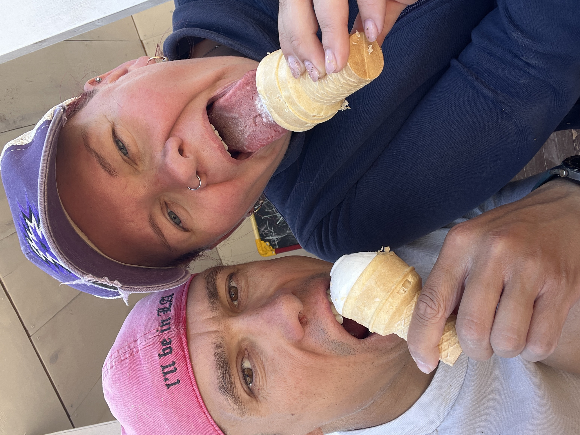 Two people, a woman and a man, are positioned in the frame with their faces close together, as if taking a selfie. The woman, wearing a purple cap, is on top and is licking the bottom of an ice cream cone. The man, wearing a pink cap with the words "I'll be in LA" written on it, is below her and has an ice cream cone close to his mouth, as if about to take a bite. The location is the city of Chaikovsky, Russia. They are outside with what looks like a wood plank wall visible behind them.
