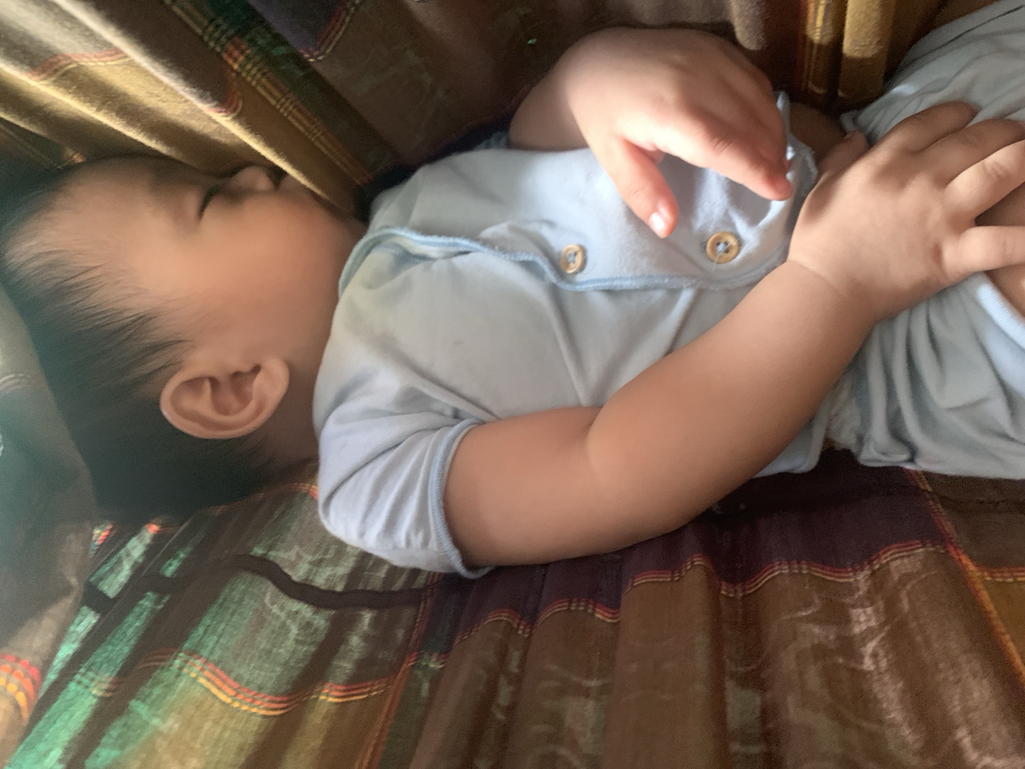 A baby is sleeping soundly in a hammock. The baby is wearing a light blue onesie with two wooden buttons. The hammock is made of fabric with a plaid pattern of brown, green, yellow, and red. The scene is likely indoors, as indicated by the soft lighting and the presence of a hammock, which is often used for napping in homes. The location in Sungai Meriam, Indonesia, is a residential area.