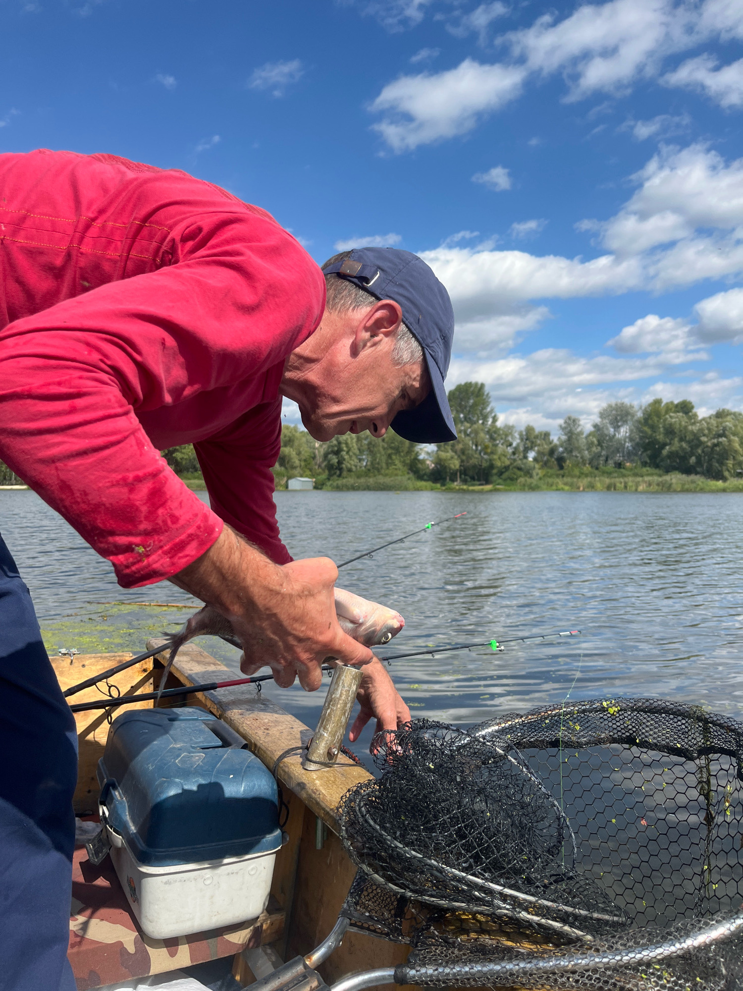 A man wearing a red long-sleeved shirt and a blue baseball cap is in a boat on a lake. He is holding a fish and appears to be removing it from a fishing rod. A blue and white tackle box is next to him, and a fishing net is in the foreground. The background shows trees and a blue sky with white clouds. The image appears to have been taken in Vyshenʹky 2, Ukraine, during daytime.