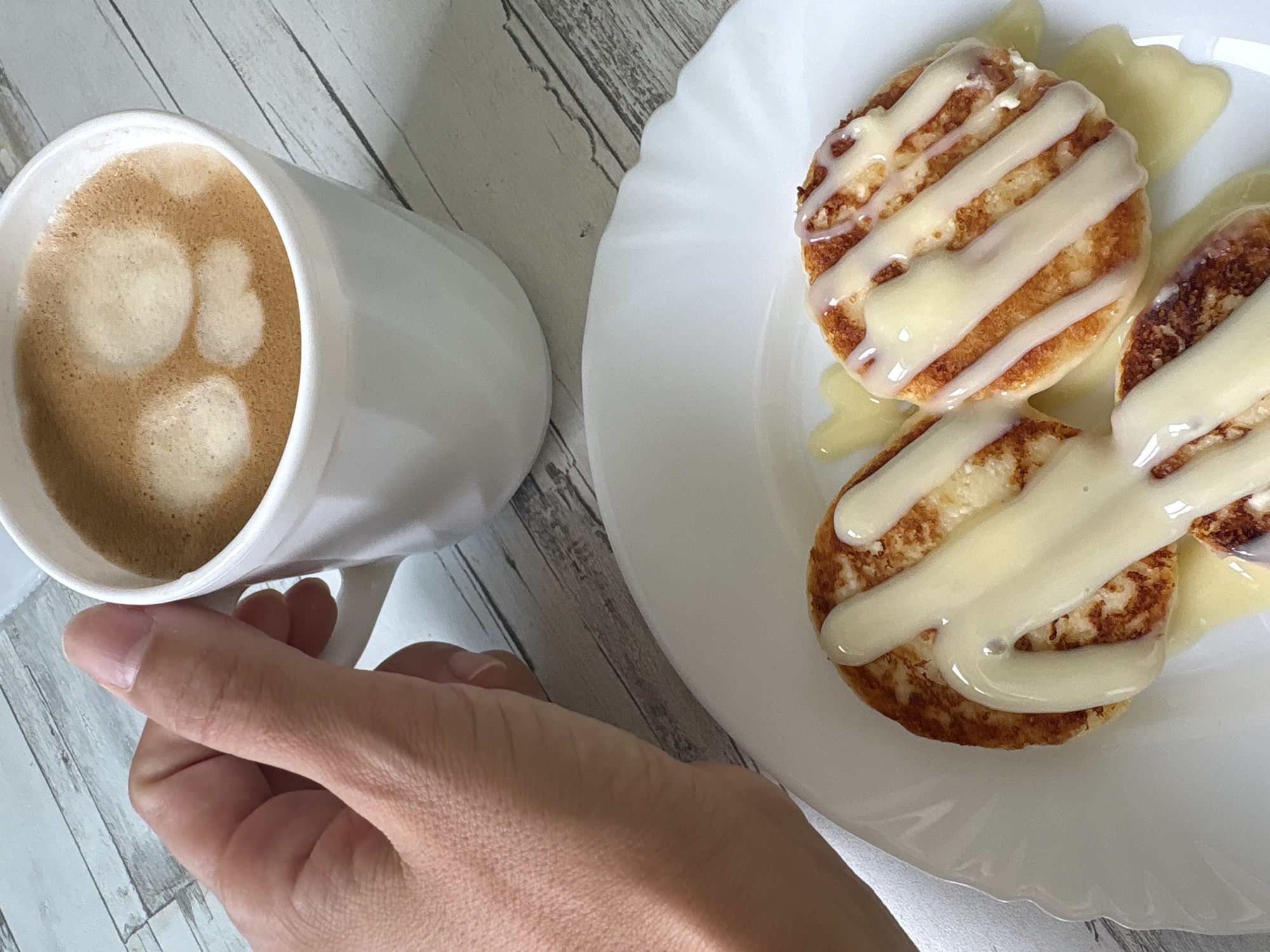 A hand holds a white ceramic mug of coffee with froth. In front of the mug, on a white plate, are two small, golden-brown pancakes drizzled with a creamy white sauce. The scene is set on a light-colored, wooden surface with a plank pattern. The overall impression is of a breakfast or snack setting. There is no identifiable information to determine the location in Chaikovsky, Russia.