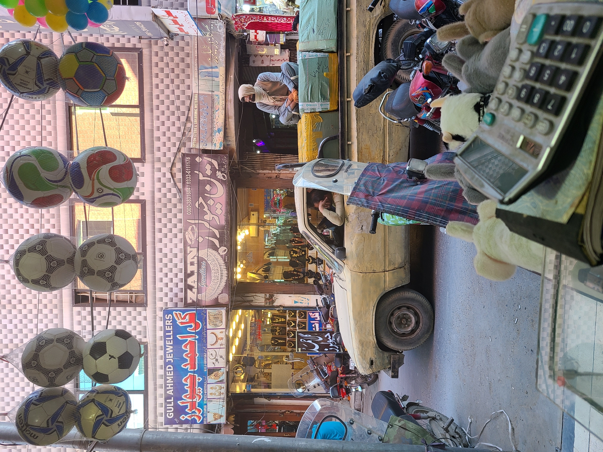 Here is a detailed description of the image:
**Location:** The image shows a busy street in an urban area, likely a commercial district in an unidentified city. Visible storefronts with signs in Urdu suggest the location is in Pakistan.
**Environment:** The scene is crowded and bustling. Overhead, strings of colorful balloons are visible. Street is narrow, with pedestrians and motorcycles. The lighting suggests daytime.
**Subjects & Objects:**
* **Street Vendor/Shop:** The viewpoint appears to be from a shop that is selling stuffed animals, calculators, and more.
* **Motorcycles:** Several motorcycles are parked along the side of the road.
* **Vehicle:** A white pickup truck is visible in the street with a person behind the wheel. Another man is on the back sitting amongst the cargo.
* **Footballs:** Strings of footballs hang near the shop.
* **Storefronts:** Several storefronts are visible, including a jewelry store ("GULL AHMED JEWELLERS").
* **Signage:** Shop signs are in Urdu, indicating the local language.
**People:** People can be seen in the vehicle, as well as on the back of it.
**Actions & Interactions:** The presence of motorcycles and the delivery truck indicate active commercial activities.
**Overall:** The image shows a lively street scene in a Pakistani city. The presence of various commercial activities, along with pedestrians, gives an impression of a vibrant urban environment.