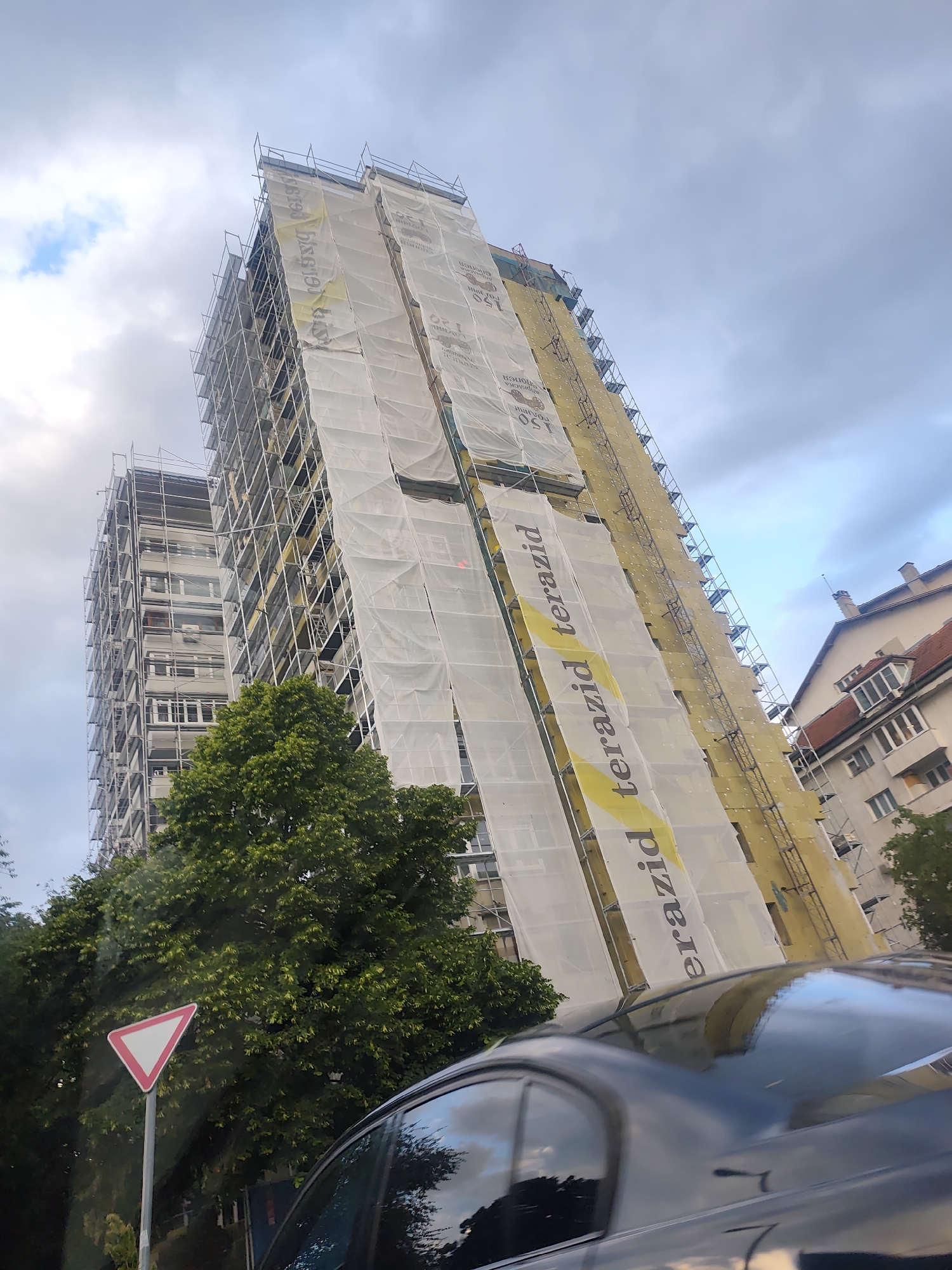 The image displays an urban scene featuring tall apartment buildings under renovation, consistent with a location like Sofia, Bulgaria. The primary subject is a multi-story building extensively covered in scaffolding. Large sections of its facade are wrapped in yellow insulation material, overlaid with white protective coverings or banners. These banners prominently display the word "terazid" in black lettering on a yellow background, oriented vertically and repeated, likely indicating a brand of construction materials or services.
To the left, a second, similarly tall building is partially visible, also surrounded by scaffolding, suggesting ongoing renovation work on multiple structures. Lush green trees occupy the mid-ground, partially obscuring the lower parts of the buildings. In the immediate foreground, the dark, reflective roof and rear window of a car are visible, indicating the photograph was taken from inside or very close to a vehicle. A white and red triangular road sign, signifying "yield" or "give way," is positioned to the left of the car and in front of the trees.
The sky is overcast with a mix of light and darker clouds, suggesting a cloudy day, possibly in the late afternoon or early evening. No individuals are visible in the scene. The architecture of the buildings, including an older structure with a red tiled roof and chimneys visible on the far right, provides contextual cues for an Eastern European city.