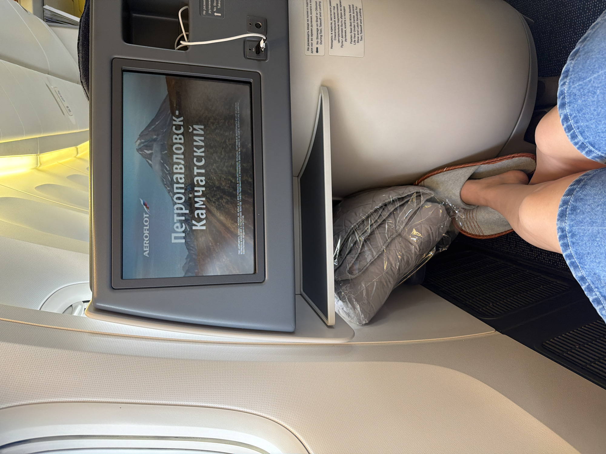 This image captures a first-person perspective from inside an airplane cabin. The primary focus is on the in-flight entertainment screen, which displays a scenic view of a mountainous landscape with the text "Петропавловск-Камчатский" and the Aeroflot logo. To the right of the screen, a person's feet are visible, wearing comfortable slippers, tucked next to a folded blanket wrapped in plastic. The person is wearing blue denim shorts, suggesting a casual travel experience.
The setting is clearly an airplane interior, indicated by the textured seats, overhead compartments, and the characteristic lighting. The presence of safety instructions labeled in both Russian and English on the wall behind the screen, mentioning "Taxi, Takeoff and Landing," further confirms the location as an aircraft. The text also includes warnings about not placing items on the shelf or under the footrest during critical phases of flight.
The image appears to be taken during daylight, as indicated by the bright illumination in the cabin. The overall mood is one of relaxed travel, with the person seemingly settling in for a flight. The view suggests the flight is either preparing for departure or has recently arrived, given the prominent display of the destination. The inclusion of the Russian destination "Петропавловск-Камчатский" strongly hints at the location being within or related to Russia.
