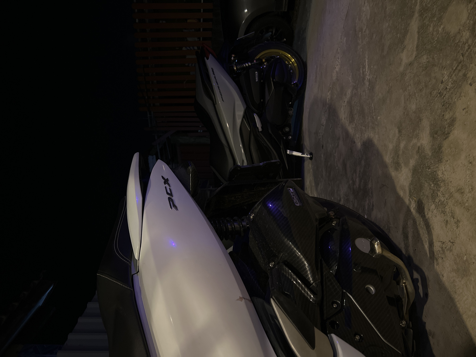 In the foreground, a white and black scooter is parked on a concrete surface, with its front end angled towards the left. The scooter features a carbon fiber accent on its lower body. Behind it, a second scooter, primarily silver and black, is also parked. This scooter has gold-rimmed wheels and a visible suspension system. The background includes a dark fence with vertical wooden slats. The lighting suggests it is nighttime or a dimly lit area. The scene is devoid of people.