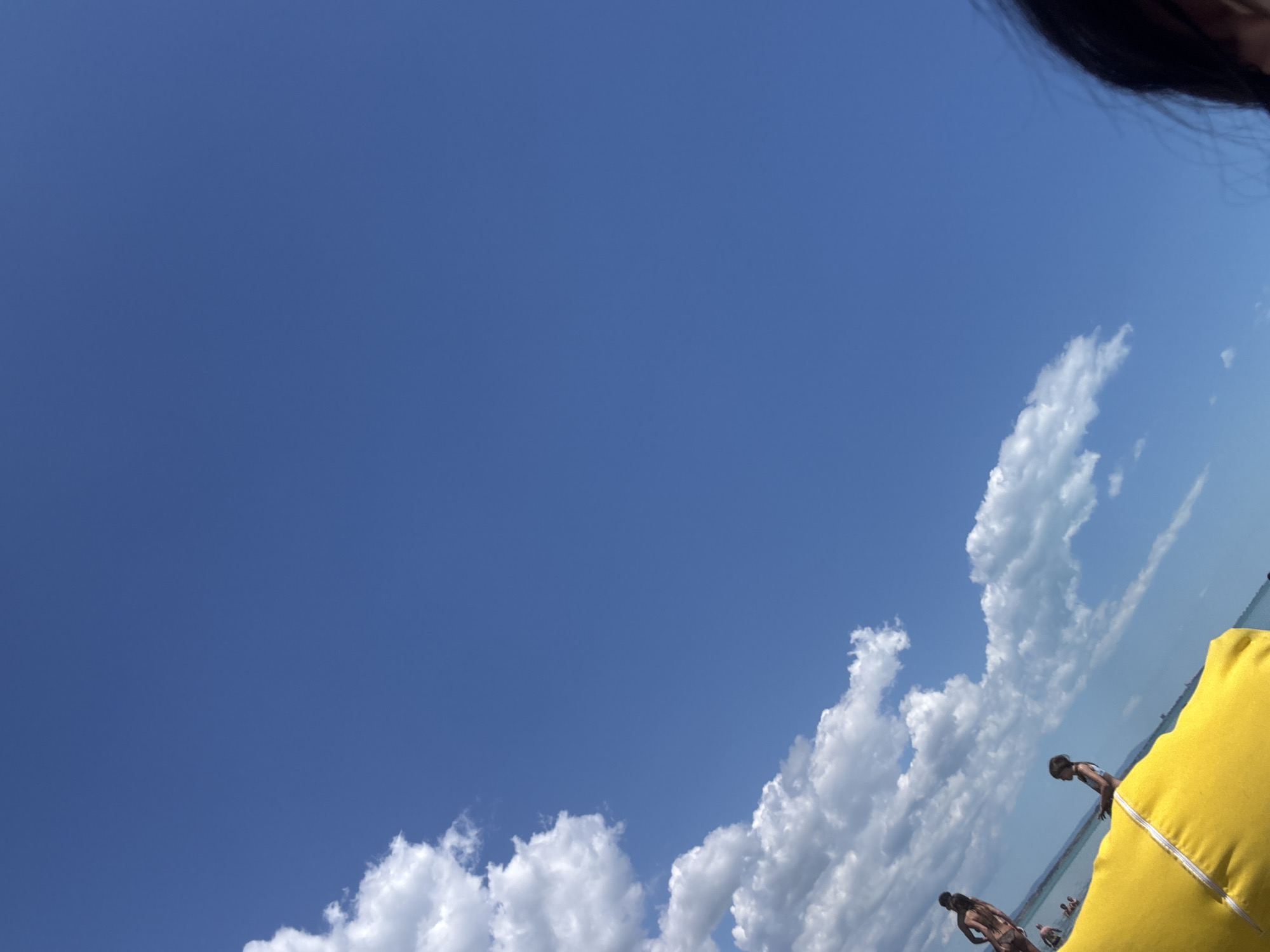 A low-angle, full outdoor shot captures a bright, clear blue sky on a sunny day, with a few fluffy white clouds scattered across the upper left portion of the frame. The scene is tilted to the right, creating a diagonal orientation. In the lower left quadrant, a bright yellow object, possibly a beach towel or cushion, is visible in the foreground. Beyond this, a body of water, likely a lake or the sea, is present. Several people are visible in and around the water. Two figures, a woman and a child, are standing in the shallow water, facing away from the camera. Further in the background, more individuals can be seen in the water. In the extreme lower right corner, the dark hair of a person is partially visible, suggesting they are also in the foreground, possibly taking the photograph. The overall impression is a relaxed, outdoor activity on a warm day, with the vast expanse of the sky dominating the scene.