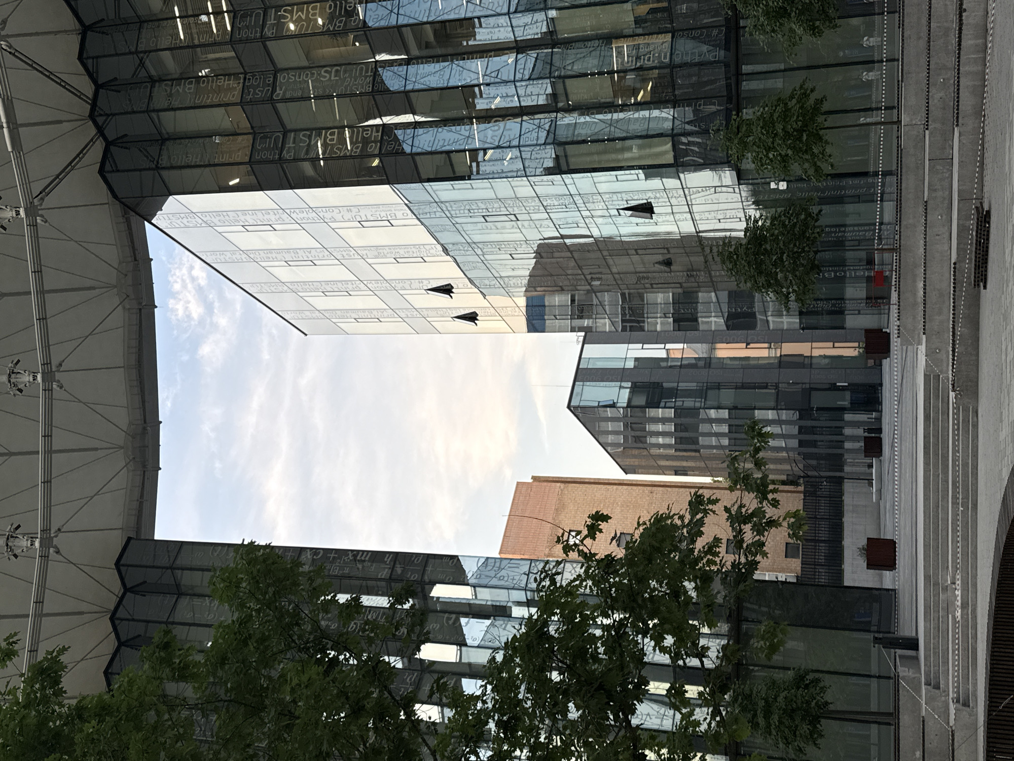 The image shows the exterior of the Moscow Institute of Physics and Technology (MIPT) campus. The central focus is on two modern glass buildings with reflective facades. The building on the right displays text inscribed on its glass panels, including programming code snippets like "Python print ('Hello World')" and "Hello BMSTU". The building on the left also has similar text and patterns on its glass. Between these two buildings, a third, older brick building is visible. The sky above is partly cloudy, with a large, white, curved architectural element spanning overhead, possibly a canopy or roof structure. In the foreground, there are concrete steps and planters with trees, suggesting an outdoor public space or plaza. The overall impression is one of a contemporary academic or research facility.