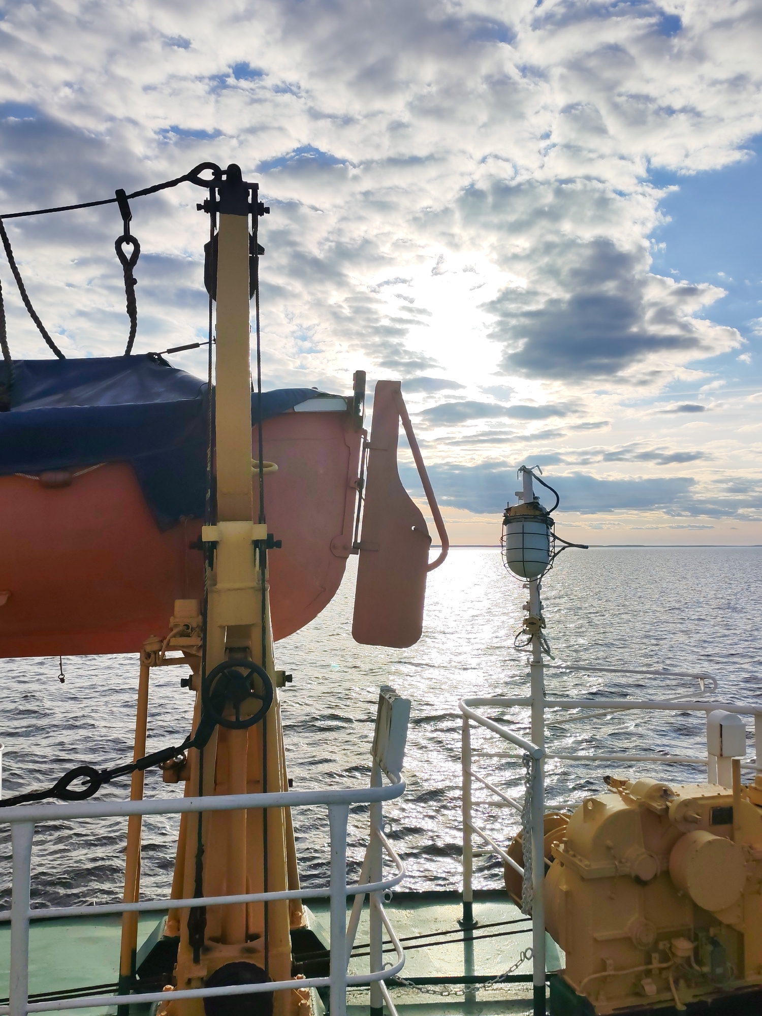 Here is a detailed description of the image:
**Scene:** The image presents a view from the deck of a vessel on the sea, under a partly cloudy sky with the sun breaking through. The location appears to be at sea, with no land visible on the horizon.
**Objects:** Prominently featured is a lifeboat with a dark blue cover, suspended on a yellow davit system. There is a safety light mounted on a pole. A white railing frames part of the lower visual field. Behind the railing, there's some yellow machinery.
**Environment:** The sky is a mixture of blue and bright clouds, with sunlight reflected on the water's surface. The sea has slight ripples.
**Weather:** The weather appears to be partly sunny.