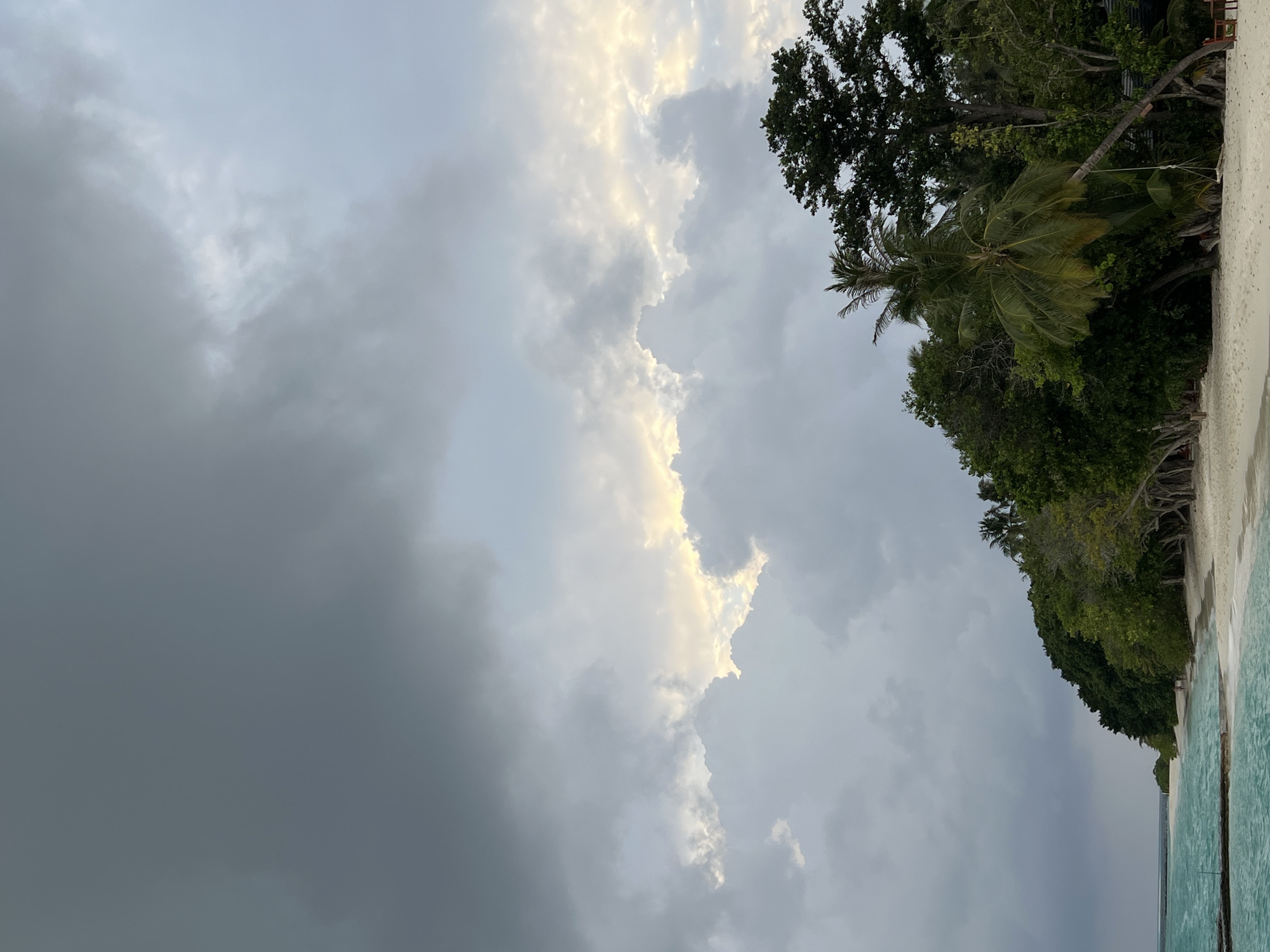 The image depicts a beach scene under a cloudy sky. The left side of the image is dominated by dark, heavy clouds, while the center shows brighter, sunlit clouds. On the right side, there's a coastline with lush green vegetation, including trees and a palm tree. The bottom right corner shows a glimpse of the ocean with a turquoise hue. The location appears to be a tropical beach, but the specific city and region are unknown.