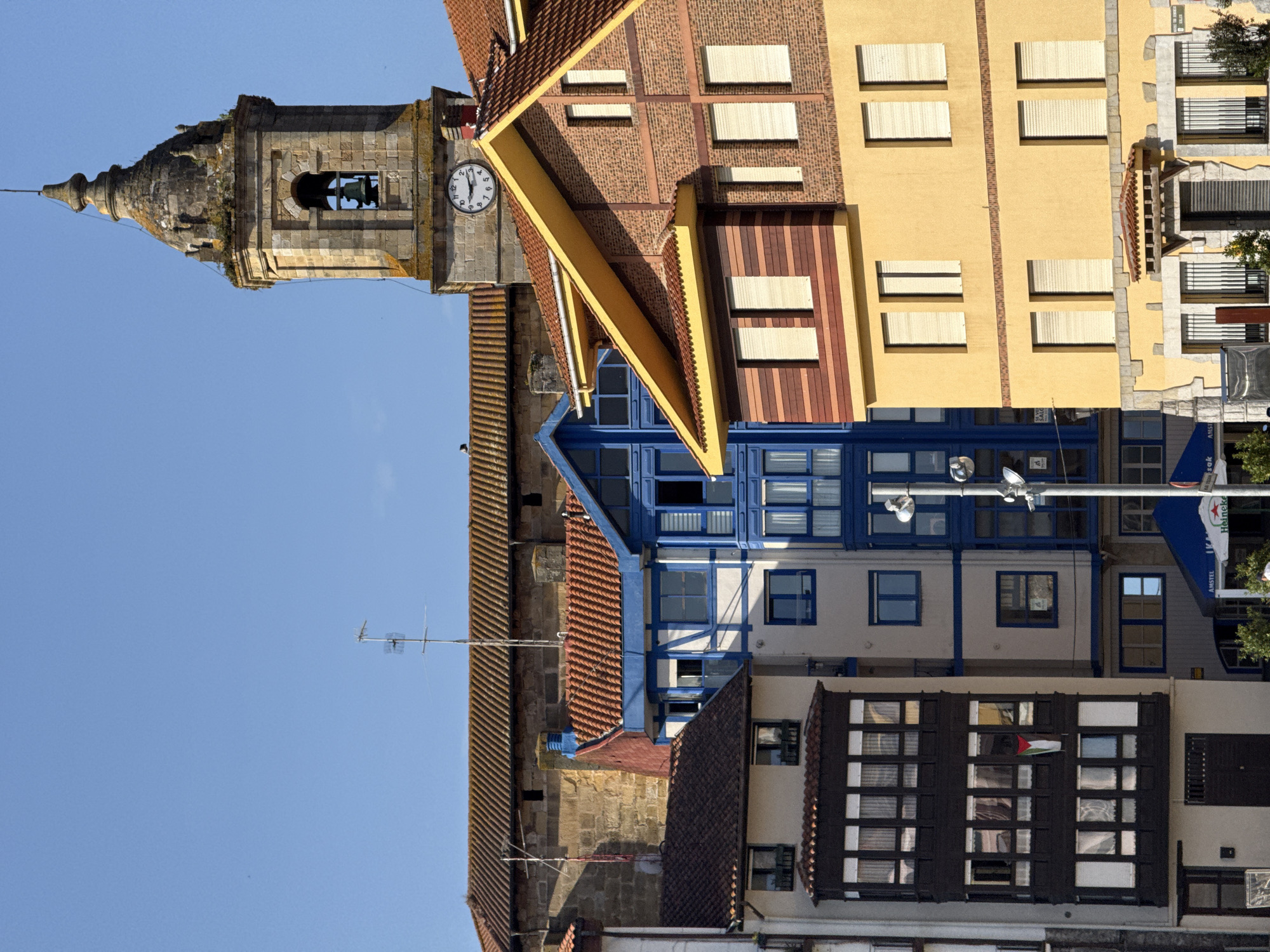 In Bermeo, Spain, a cluster of buildings is captured under a clear blue sky. The scene is dominated by the architecture, featuring a prominent church tower with a bell and a clock, its stone facade weathered and adorned with greenery. Adjacent to it, a series of multi-story buildings showcase varied architectural styles. Some exhibit traditional red-tiled roofs and white walls with blue trim, while others are clad in warm-toned brick and painted yellow.
The buildings have numerous windows, many covered by slatted blinds. One building features a dark, ornate balcony with multiple windows on its upper floors. A flag, possibly of Palestine, hangs from one of these windows. Below street level, awnings and signage for businesses like "AMSTEL" and "Heineken" are visible, suggesting a commercial area. The overall impression is one of a sunlit, urban coastal environment.