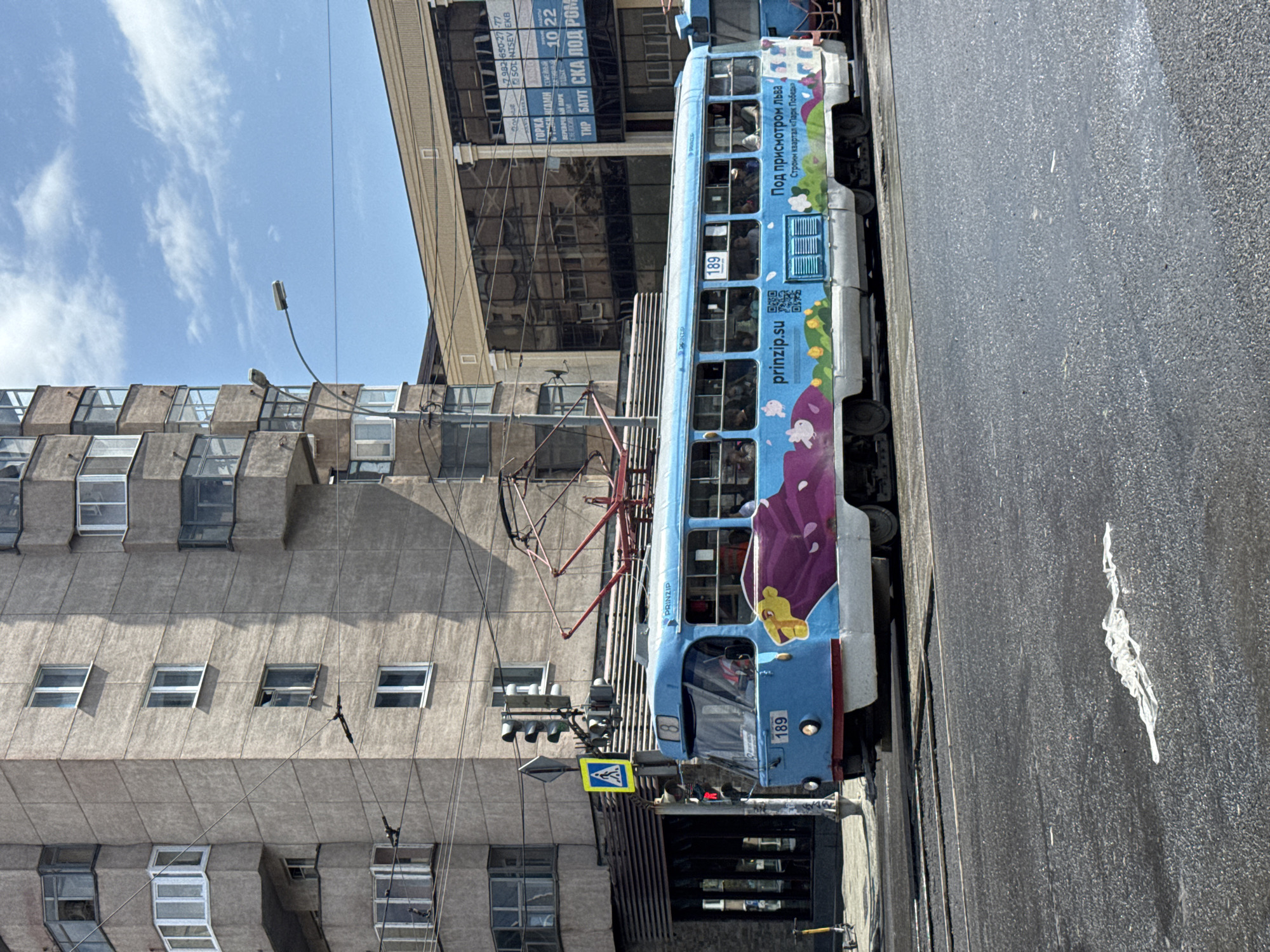 This media file shows a light blue tram traveling along a street in Yekaterinburg, Russia. The tram is decorated with a colorful advertisement featuring purple floral designs and yellow cartoon characters. The text on the tram includes "prinzip.su" and Russian phrases such as "Под присмотром льва, Строим квартал «Парк Победы»" which translates to "Under the lion's supervision, We are building the quarter 'Victory Park'." Another visible number on the tram is "189".
The tram is on a paved road, with tram tracks visible. To the left of the tram, a large, multi-story brutalist-style building with many windows dominates the scene. Above the building, a bright blue sky with wispy white clouds suggests a clear and sunny day, likely during daylight hours. A streetlamp is also visible in the upper left quadrant of the image. Traffic signals and a pedestrian crossing sign are present near the tram, indicating an urban street environment. The overall atmosphere appears to be a typical urban scene on a pleasant day.