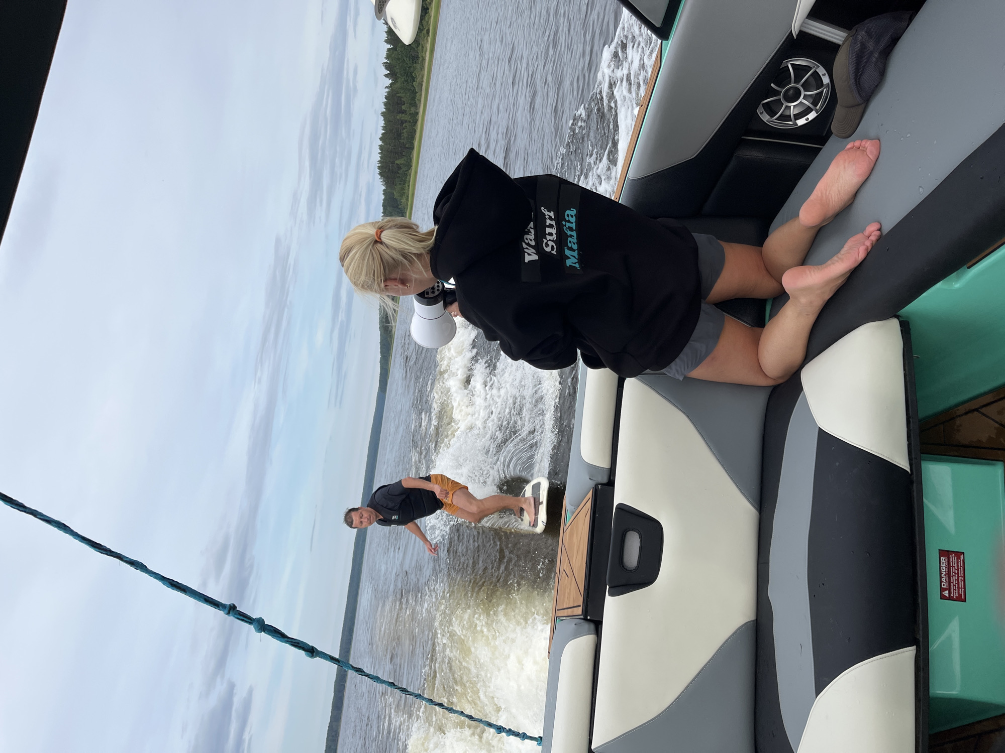An overhead view captures a man wake surfing on a lake, viewed from the back of a boat. The man, wearing a black life vest and orange shorts, is mid-action, carving a wave. In the foreground, a woman with blonde hair pulled into a ponytail kneels on the boat's upholstered seating, holding a white megaphone to her mouth. She is wearing a black hoodie with text on the back that reads "Wake Surf Mafia" and grey shorts. The boat is on a body of water, with a treed shoreline visible in the distance under a cloudy sky. The boat's interior features grey, black, and white seating. A "DANGER" warning label is visible on a green compartment. The specific location is the city of Gromovo, Russia.
