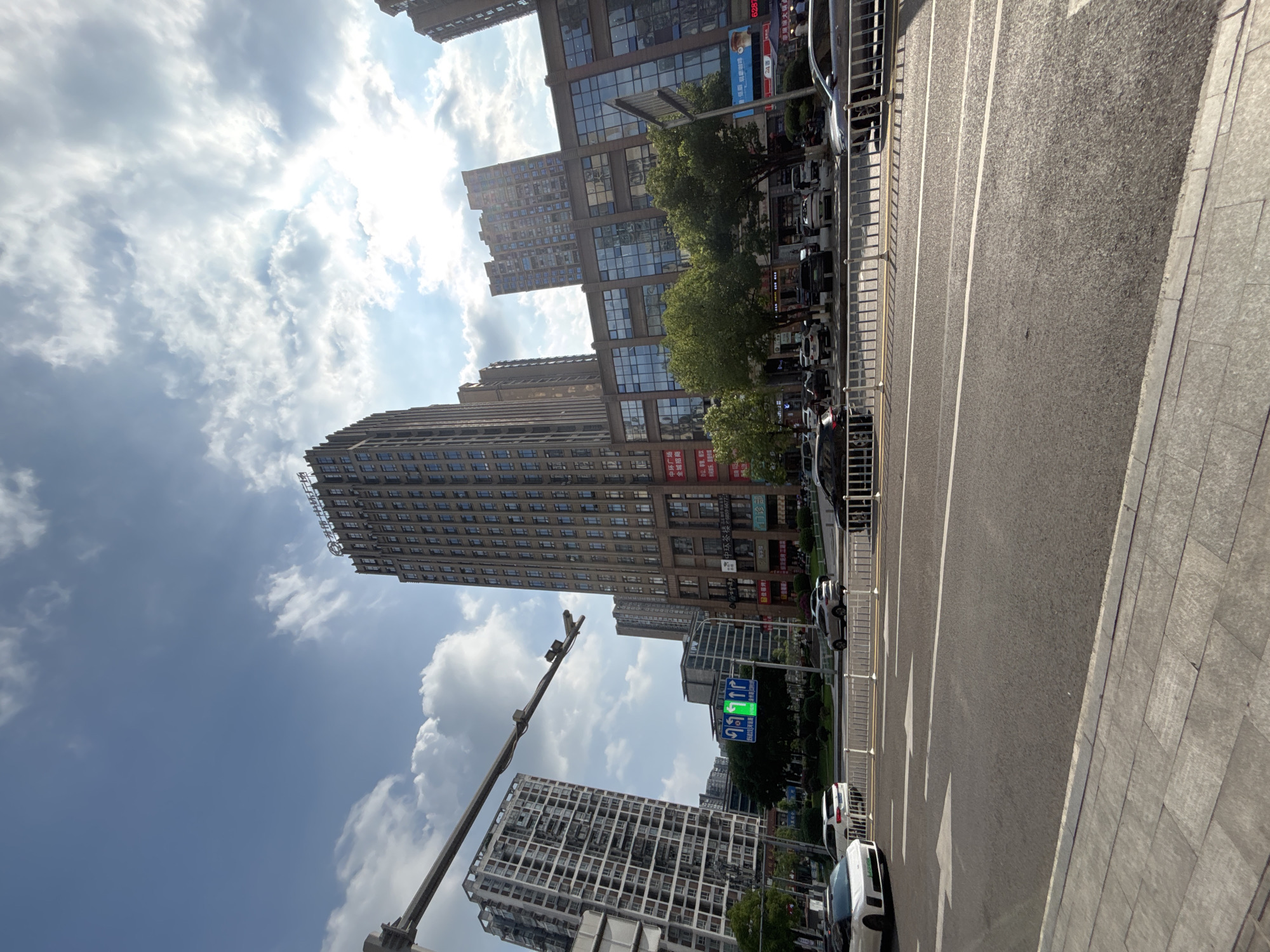 A bright, sunny day in Yuzhong District, China, with a clear blue sky dotted with white clouds. The scene depicts a busy urban street with several tall buildings dominating the skyline. In the foreground, a multi-lane road with white lane markings and arrows guides traffic. Several cars are visible, including a white sports car on the left and various SUVs and sedans further down the road. A pedestrian walkway with gray paving stones lines the road.
The middle ground features a traffic light with a green arrow indicating traffic can turn left, and other directional signs. Trees are interspersed between buildings, adding a touch of greenery to the urban landscape. The buildings themselves vary in architectural style and height, with some appearing to be modern residential apartments and others commercial structures with glass facades. One prominent building in the center has Chinese characters and logos on its exterior. The overall impression is one of a thriving, modern city on a pleasant day.