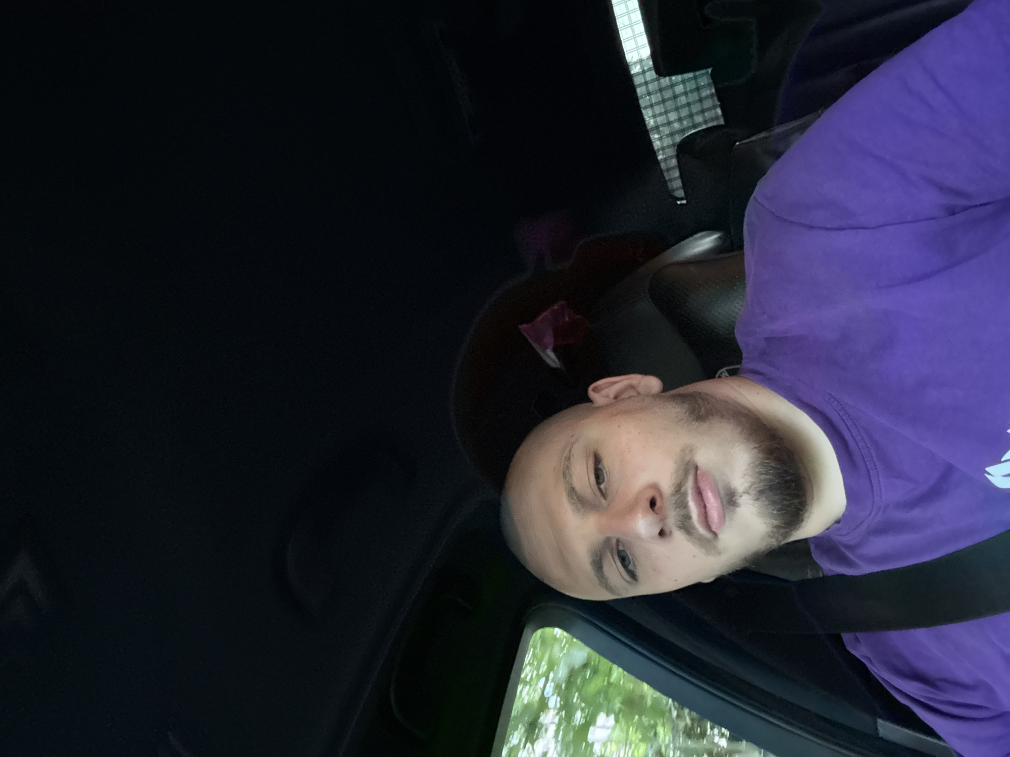 A young adult male with a shaved head and a short, dark beard is seated in the driver's seat of a vehicle in Stara Zagora, Bulgaria. He is wearing a purple crew-neck t-shirt and a black seatbelt across his chest. His light-colored eyes are directed towards the camera, maintaining a neutral expression. The car's interior features dark-colored upholstery and headliner. Through the driver's side window, blurred green foliage is visible, suggesting movement or proximity to trees. In the rear of the vehicle, a dark headrest is visible, and a grid-like pattern can be seen through the back window, possibly a cargo net or window tint. A small, reddish-purple fabric object is partially visible behind the driver's head on the seat.