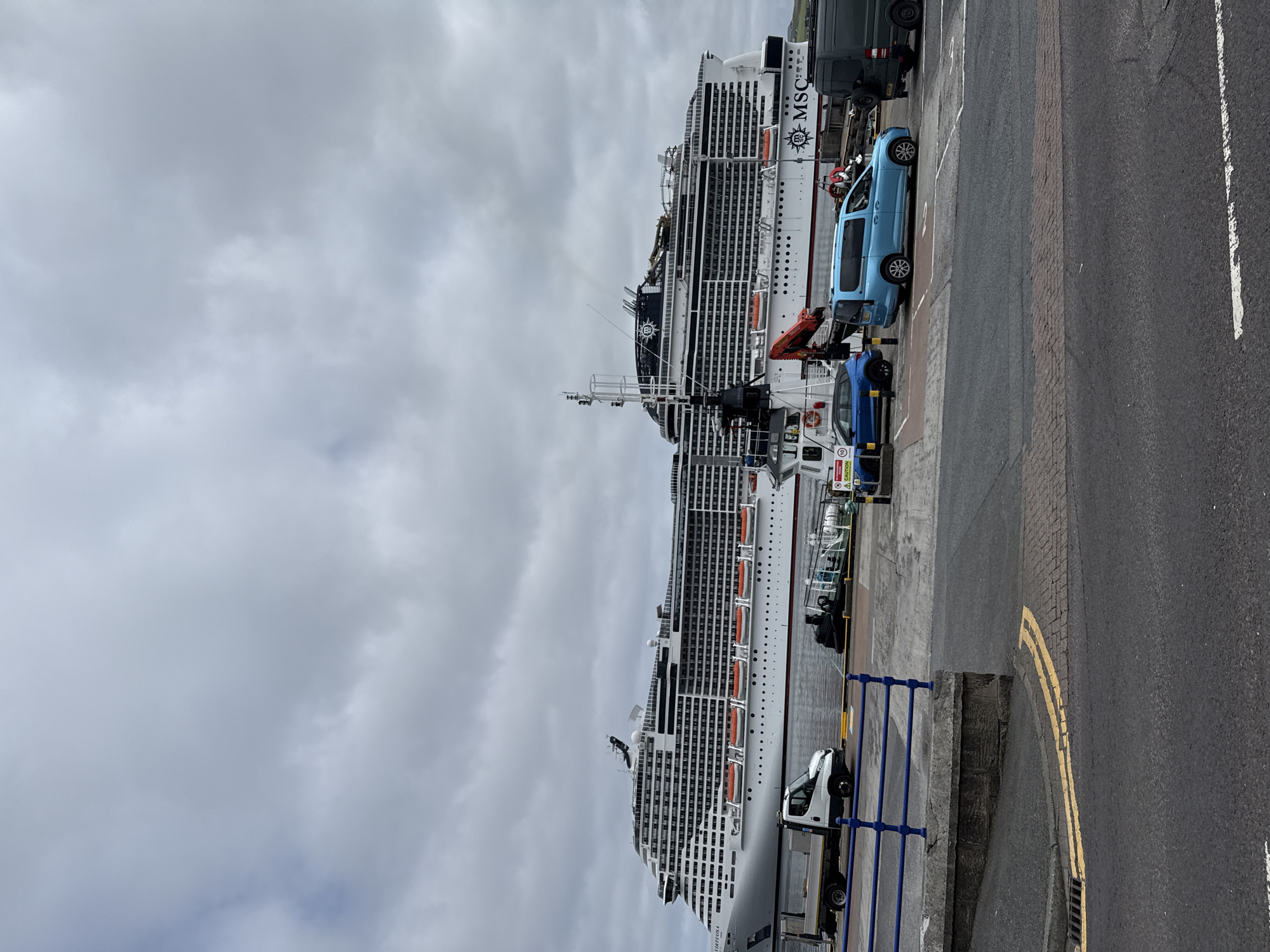 A large white cruise ship, identified by the "MSC" logo, is docked at a port under a cloudy sky. The ship, named "Virtuosa" on its side, is positioned parallel to the quay. In the foreground, a paved road with double yellow lines runs horizontally. To the left of the road, a blue metal railing separates the walkway from the port area. Several vehicles are visible on the quay, including a white truck with a flatbed, a blue car, a light blue van, and a dark colored van. A red crane is attached to a structure on the ship. The environment appears to be a commercial port. The location is the port of Lerwick, United Kingdom, which is a hub for cruise ships and cargo vessels in the Shetland Islands.