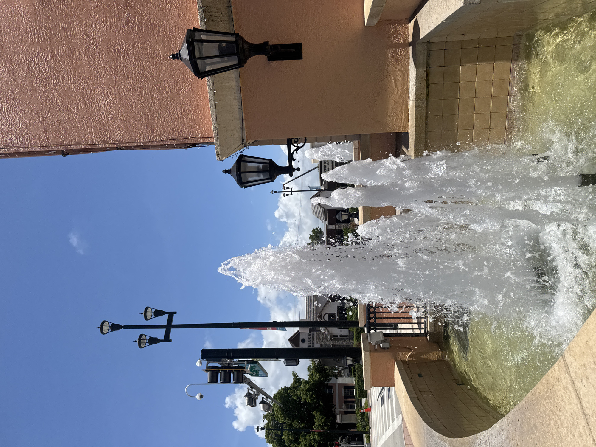 This is a medium shot, eye-level, outdoor photograph of a decorative fountain located in front of a building with an orange stucco exterior in Boca Raton, Florida, United States of America. The fountain has multiple water jets spraying upwards into the clear, blue sky. The water is a pale green color. The fountain is made of beige stone and has a black metal fence behind it. The building's exterior is textured orange stucco with a grey stone trim. Two black, ornate street lamps are mounted on the building's exterior. Another black street lamp with three lights is visible in the background. A traffic light and a security camera are also visible in the background, along with trees and other buildings. The sun is shining brightly, casting shadows.