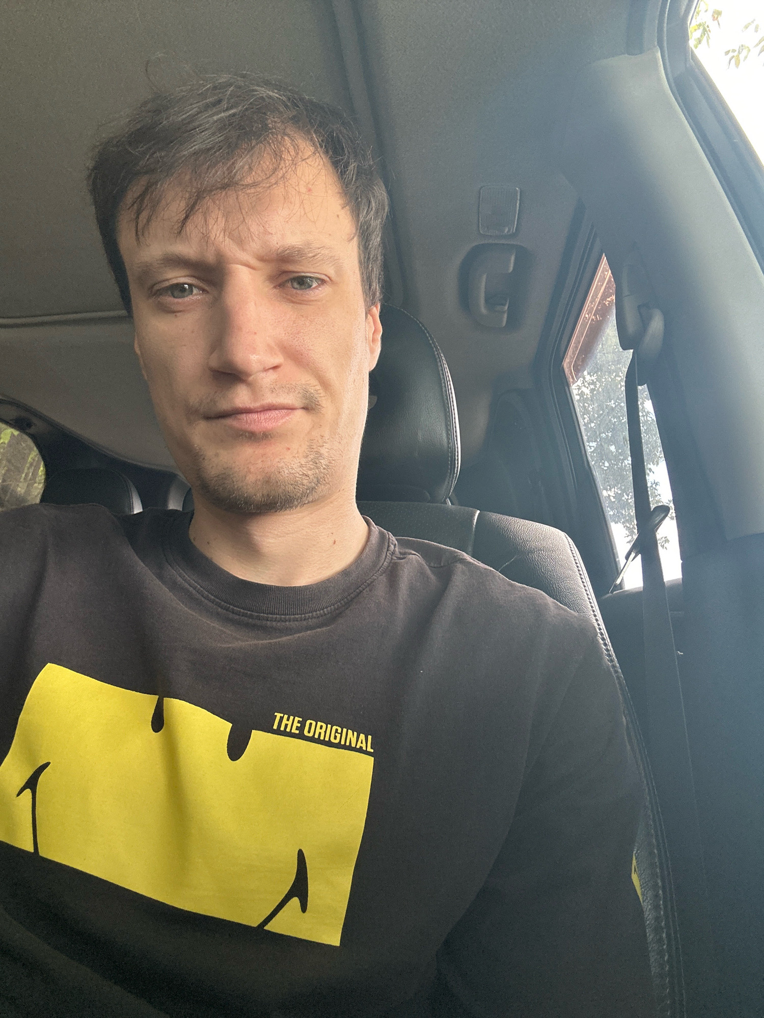 A man with short brown hair and a stubble beard is sitting in the driver's seat of a car, looking at the camera with a slight frown. He is wearing a black t-shirt with a yellow smiley face graphic and the text "THE ORIGINAL" above it. The car interior is visible, including the headrest of the passenger seat and a part of the dashboard. Sunlight streams in through the window on the right, illuminating the scene. The background suggests an outdoor environment with trees visible through the car window. The image appears to be a selfie taken inside a vehicle in Makiv, Ukraine.