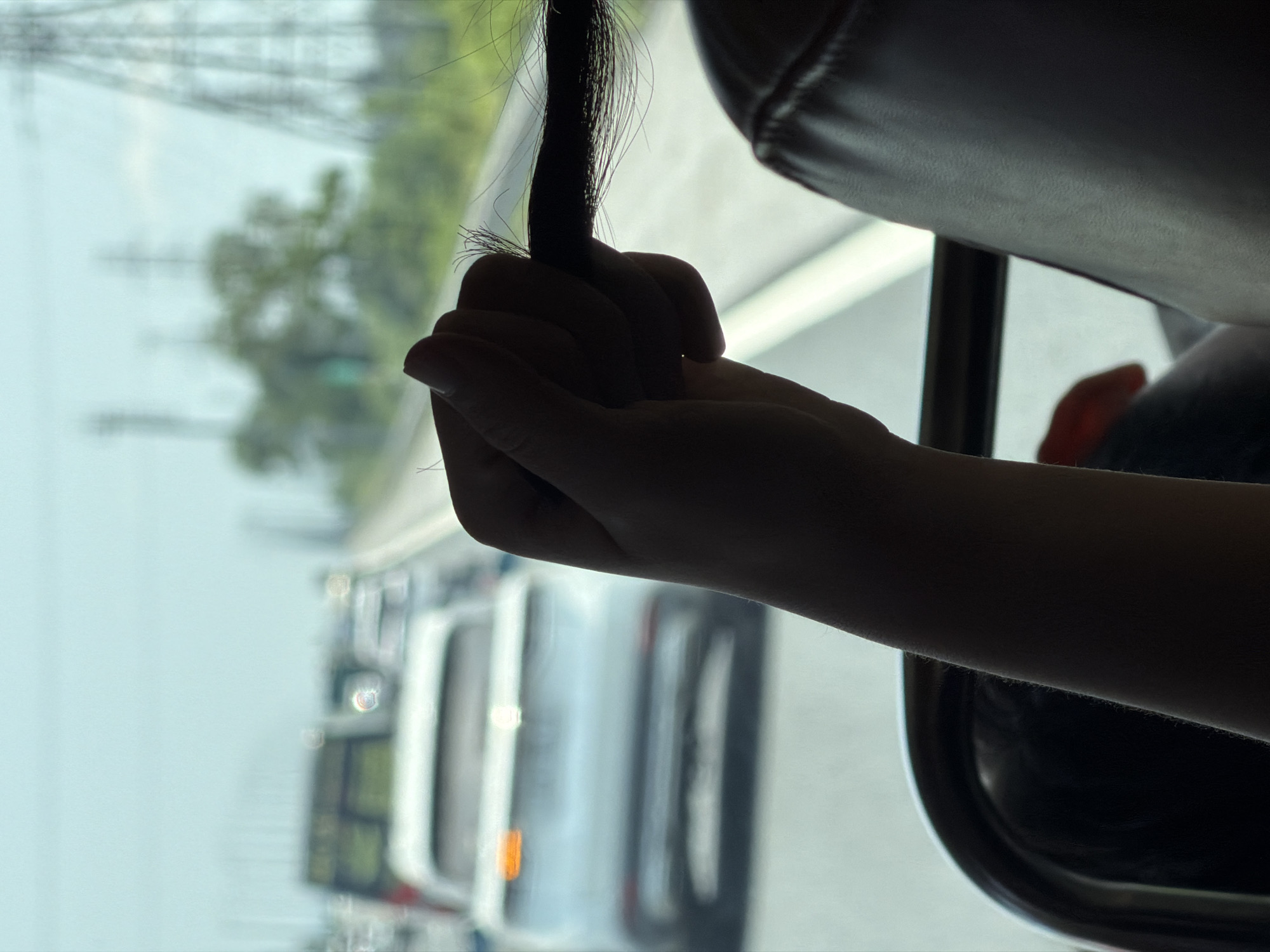 A hand is clutching a tuft of black hair inside a moving vehicle. The window shows a blurred cityscape with vehicles. While it's hard to pinpoint the exact location as Hải Phòng, Vietnam without additional information, the presence of vans and the general landscape suggest an urban setting. The photo seems to have been taken from the back seat of a vehicle, with a headrest visible in the top right.