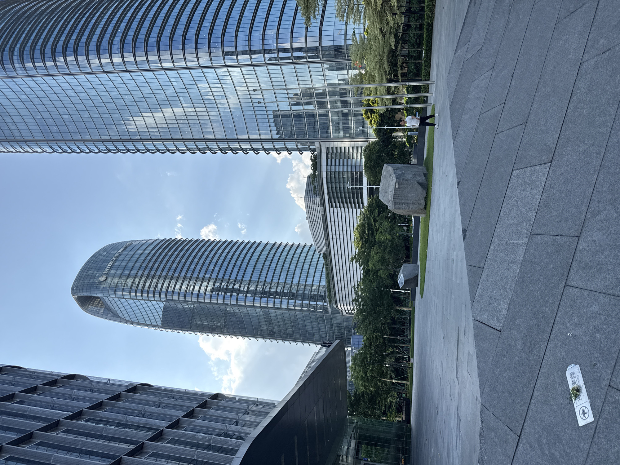 The image depicts the exterior of a modern urban environment, likely a business or commercial district, in Haizhu District, China. In the foreground, a gray, textured stone plaza is visible, with a few large rocks and some scattered greenery. In the mid-ground, a person is walking across the plaza. The background is dominated by several tall, contemporary glass skyscrapers, their facades reflecting the blue sky and scattered clouds. One prominent building, the InterContinental Hotel Guangzhou Exhibition Center, is visible with its distinctive curved shape and the hotel's logo. The architecture suggests a well-developed, possibly recently constructed, area with an emphasis on modern design and materials.