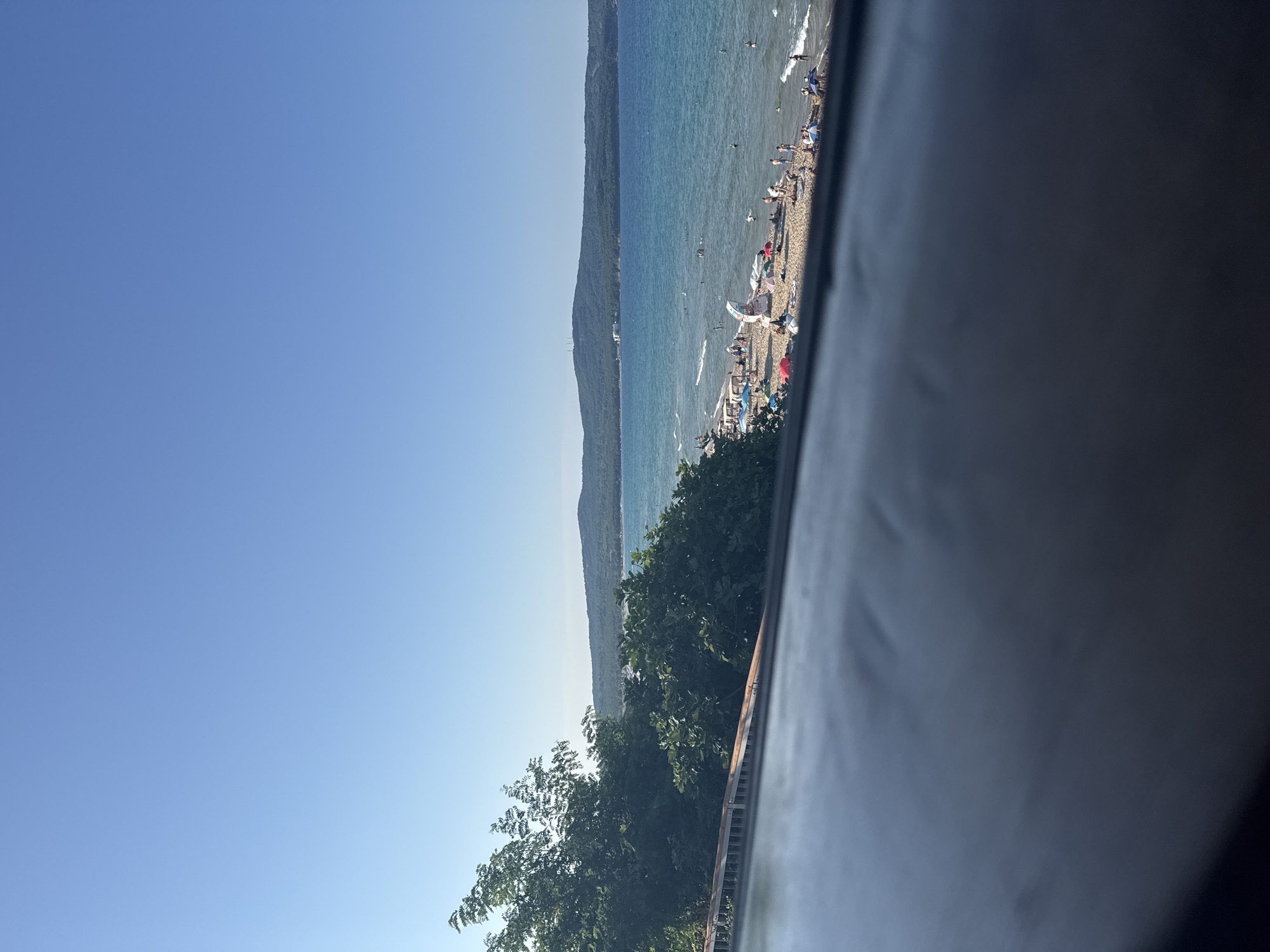 The image captures a bright, sunny day at the beach in Primorsko, Bulgaria. The foreground is dominated by a dark, out-of-focus surface, possibly a railing or car interior, obscuring the bottom part of the scene. Beyond this, lush green trees line the left side of the image, with their foliage extending towards the sea.
The main focus is the beach area, filled with people enjoying the warm weather. Numerous beach umbrellas and tents are scattered across the sand, providing shade for sunbathers. Several individuals are visible walking, sitting, and lying on the beach. Children can be seen playing near the water's edge.
The sea is a vibrant turquoise, with gentle waves lapping against the sandy shore. Some people are swimming or wading in the water, while others are further out. A few boats are dotted on the horizon.
In the background, a large, forested hill rises from the coastline, its slopes covered in dense green vegetation. The sky is a clear, cloudless blue. The overall impression is of a busy and lively beach scene, characteristic of a popular summer tourist destination.