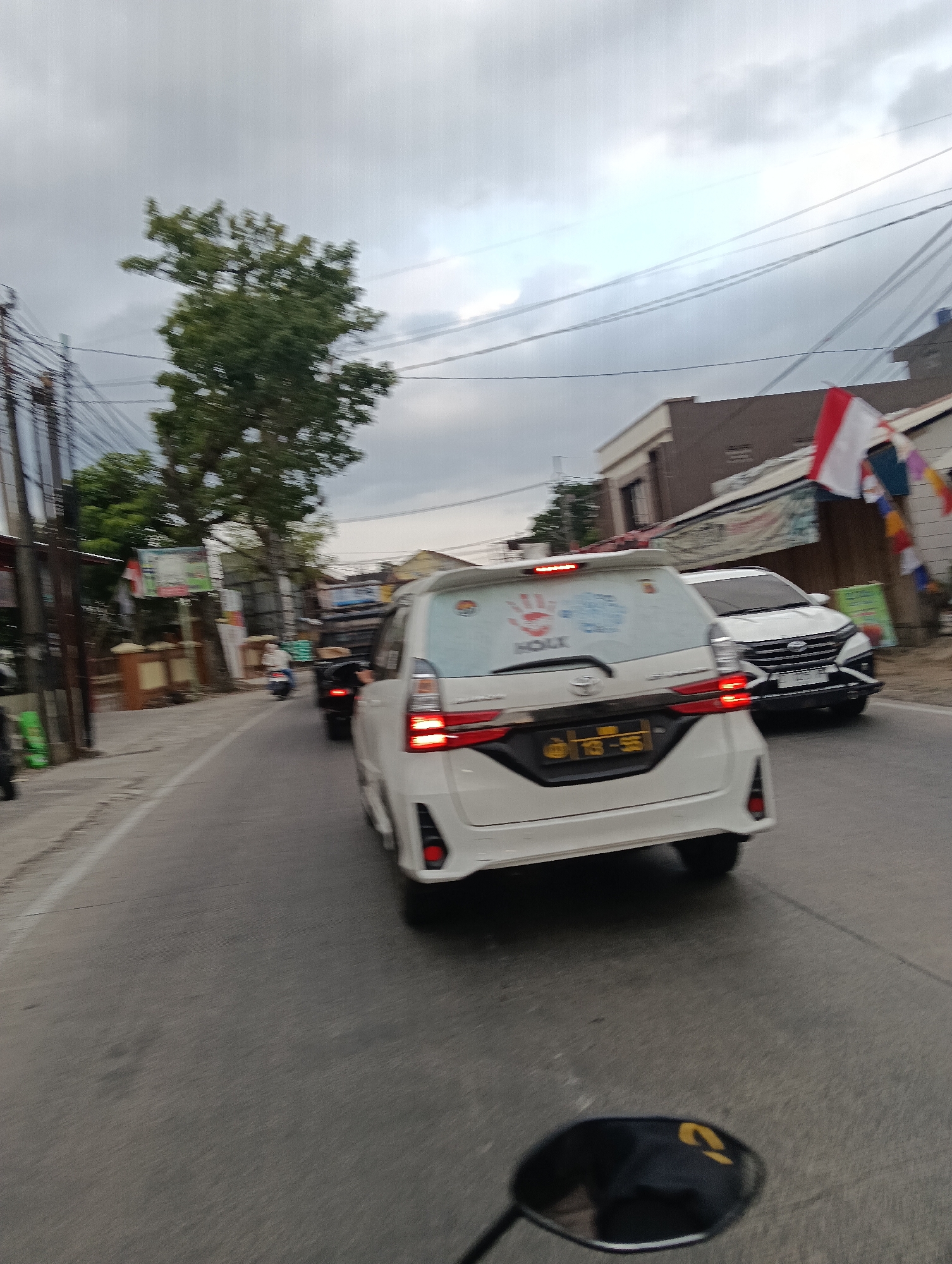 The image shows a view from a motorcycle traveling on a paved road in Desa Cipakat, Indonesia. A white MPV is in the foreground, with its rear visible. The car has a license plate that reads "138 - SE". There is a red handprint graphic on the rear window of the MPV, with text beneath it. To the right of the MPV, a white SUV is driving in the same direction. In the background, there are buildings, trees, utility poles with many wires, and a few Indonesian flags are visible, suggesting a festive occasion. The sky is overcast. Another motorcycle is visible further up the road. The scene depicts a typical street in a residential or commercial area during daytime, possibly with traffic.