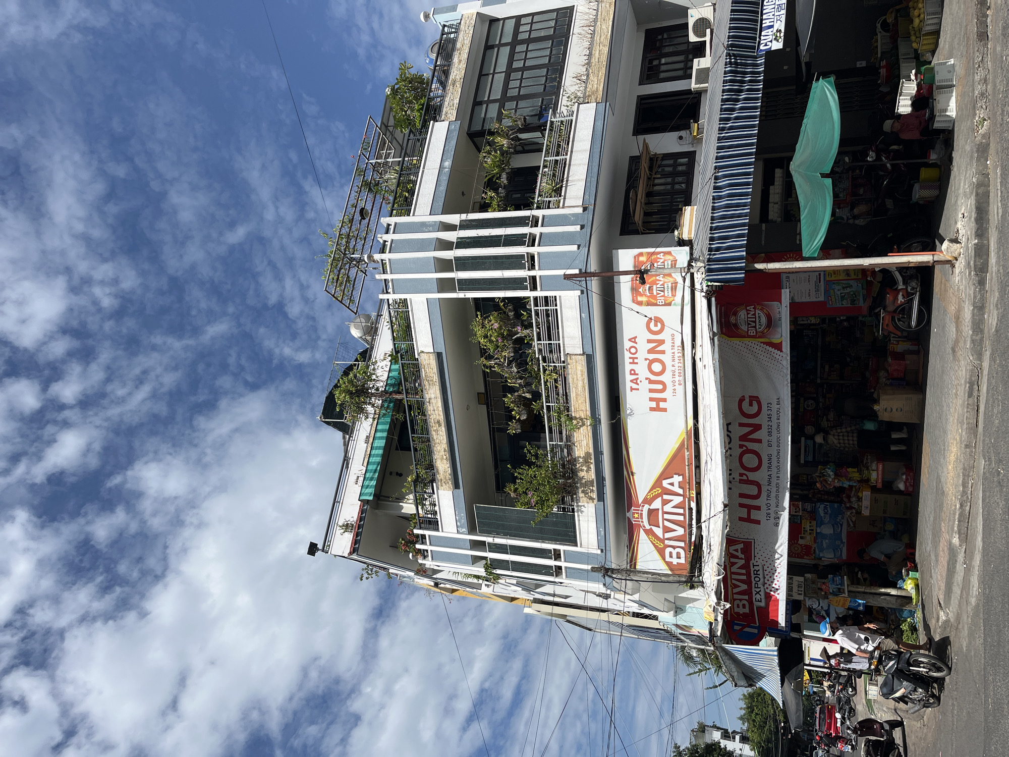The image shows a street view in Nha Trang, Vietnam, featuring a multi-story building housing a store. The building has a modern facade with balconies adorned with plants. The ground floor is occupied by a shop with awnings and displays of goods, including drinks and produce. Several motorcycles are parked on the street. People are visible in and around the store, some appearing to be customers or vendors. The sky is blue with scattered clouds. A banner on the building reads "TẠP HÓA HƯƠNG" and includes the address "126 VÕ TRƯỜNG, P. NHA TRANG".