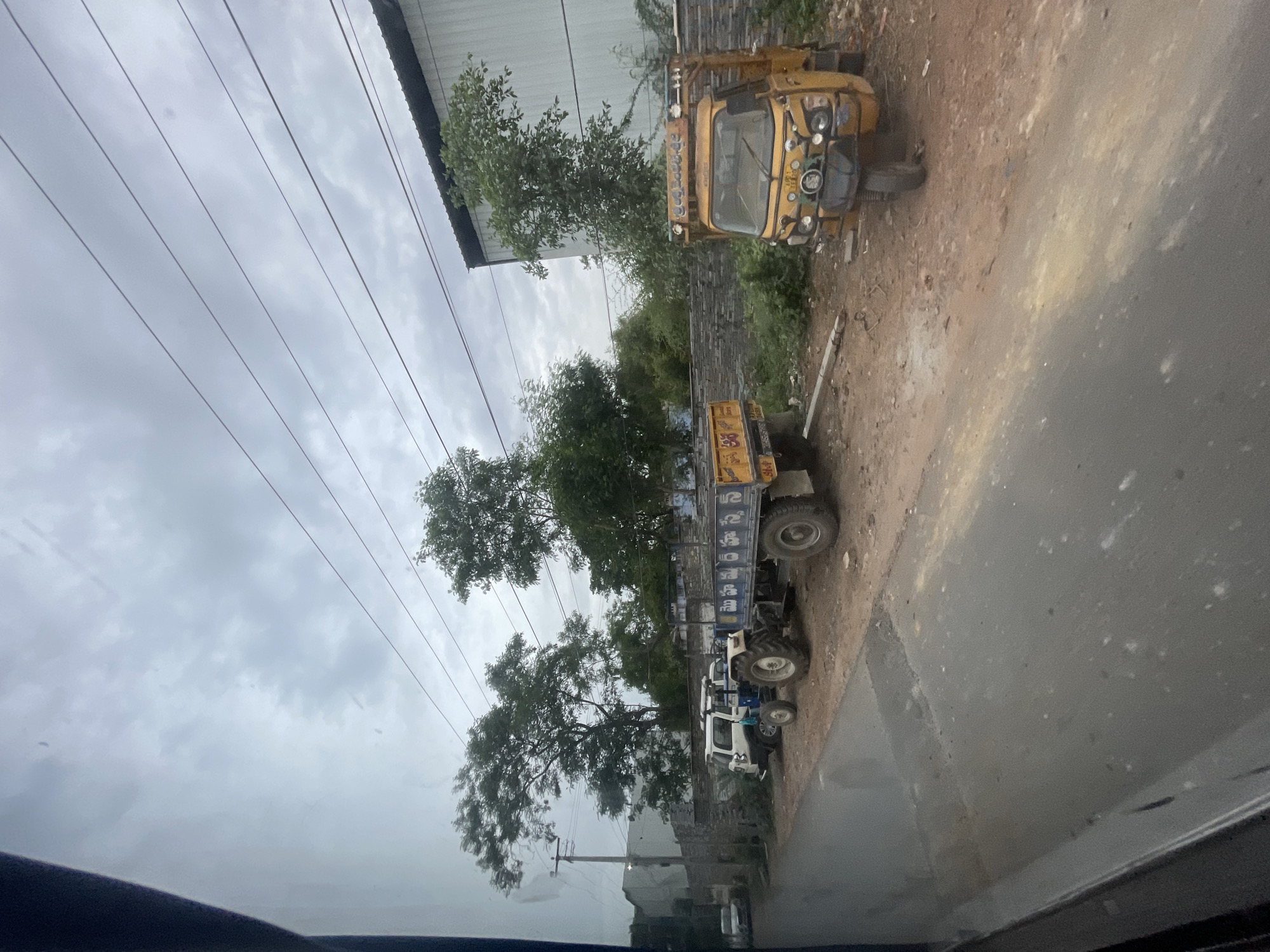The image depicts a street scene in Bethamcherla, India, under an overcast sky. On the right side of the road, a yellow three-wheeled auto-rickshaw is parked. Adjacent to it, a blue and yellow tractor with a trailer is stopped. To the left of the tractor, a white SUV is parked. In the background, a long, low building with a dark roof is visible, likely an industrial or commercial structure. Trees are scattered throughout the scene, with denser foliage behind a low stone wall separating the road from the background structures. Power lines are strung across the sky. The road itself is paved, with a dirt and gravel area alongside where the vehicles are parked. The overall impression is of a functional, working area within the town.