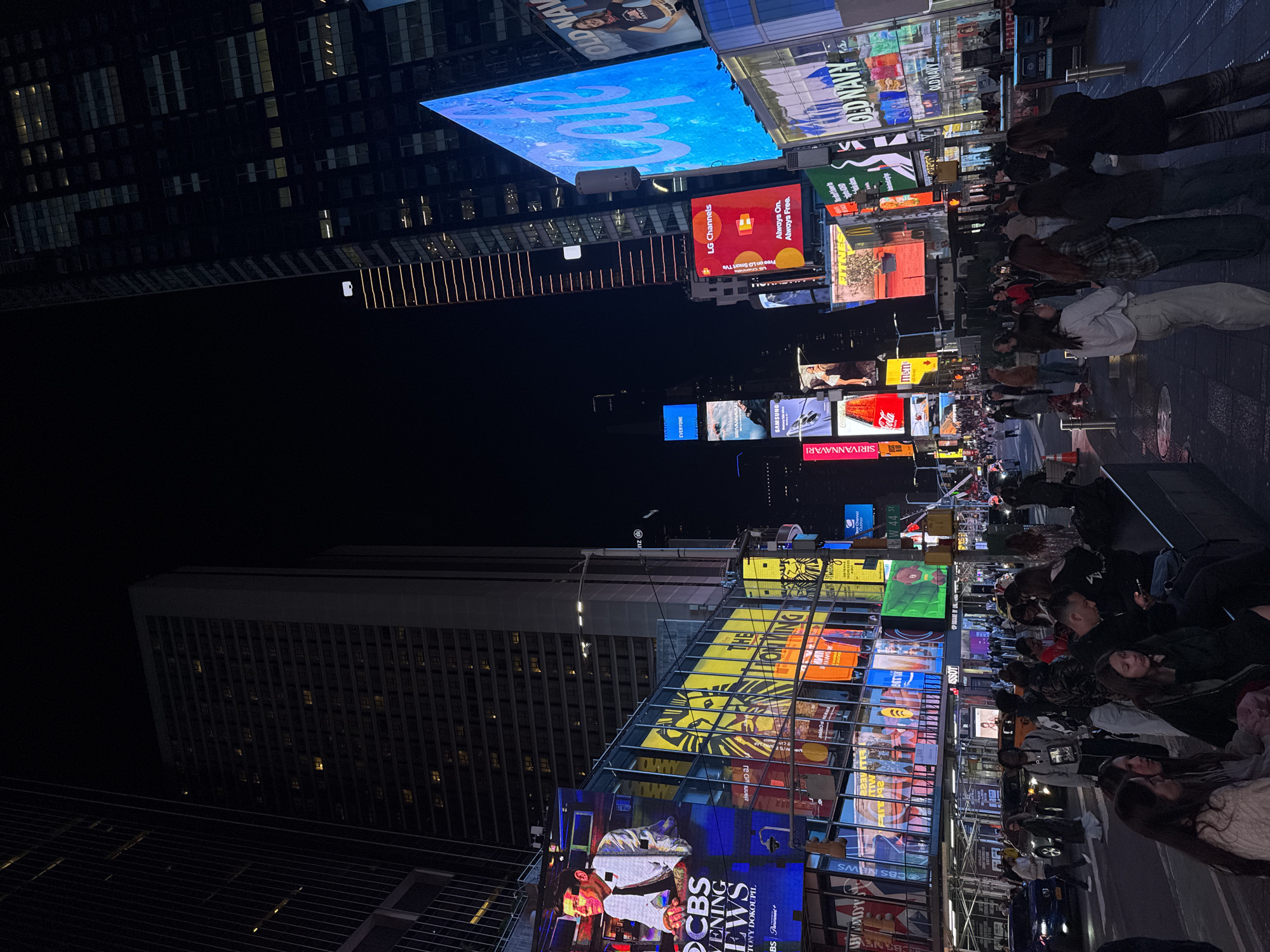 This image shows a nighttime view of Times Square in New York City. The scene is dominated by large electronic billboards displaying various advertisements and media content. Pedestrians are visible, walking or standing, their faces lit by the light from the billboards. The skyscrapers in the background are visible, with some of their windows illuminated. The overall impression is that of a crowded urban environment, characterized by bright lights and the energy of a popular tourist destination.