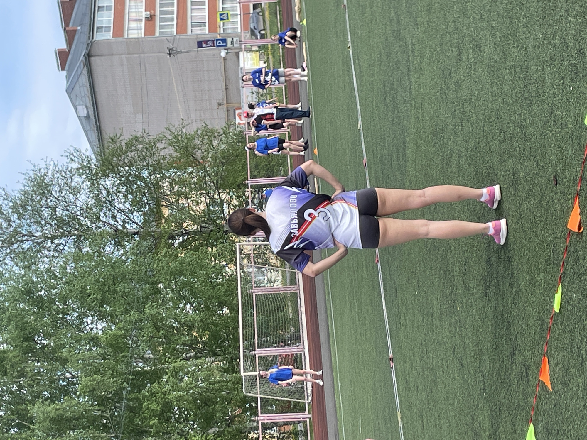 The image shows a group of people on a green field in Zav'yalovo, Russia. There is a woman in the foreground, wearing a white and purple sports jersey with the name "ZAVYALOVA" printed on the back. She is standing with her hands on her hips. Behind her, there are other people dressed in sports uniforms lined up. A building is in the background. There are green trees visible. The field has white lines and orange and green triangular flags. A goal net structure is also visible.