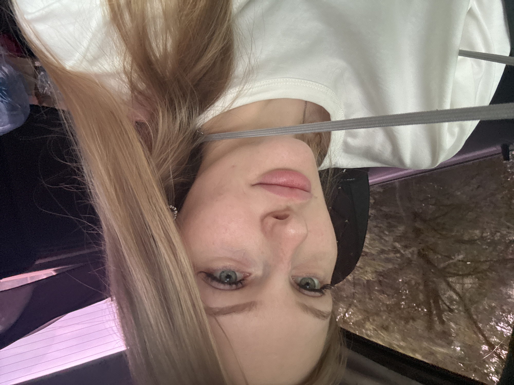 A young woman with blonde hair and blue eyes is looking forward while sitting in a car. She is wearing a white t-shirt. The background outside the car window shows trees. The woman is likely in a natural setting or on the outskirts of Perm, Russia.