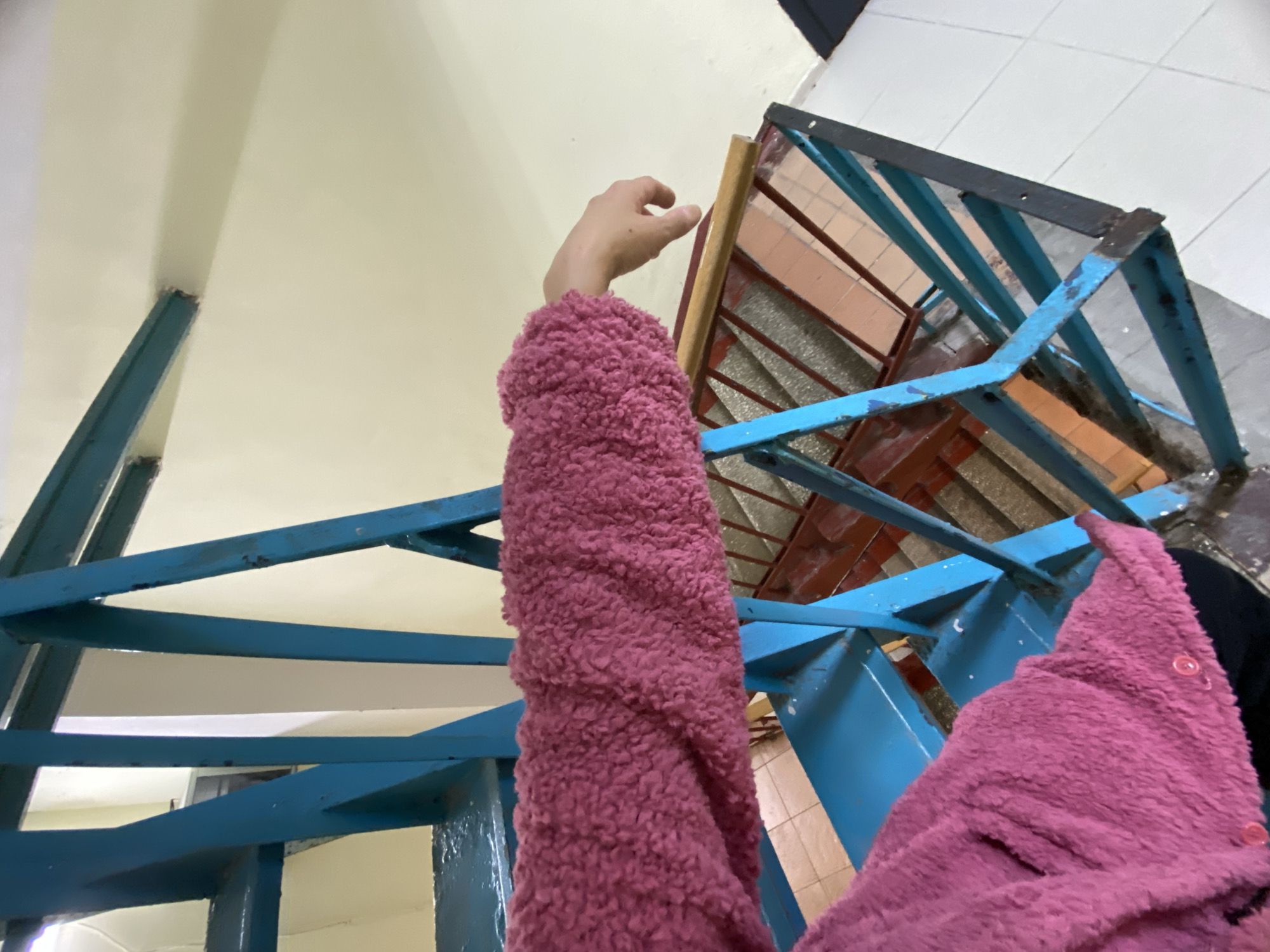 The image presents an upward-angled view within what appears to be an indoor building space, likely a stairwell or corridor in Moscow, Russia. A person is visible only through their right arm and a portion of their torso, which is covered by a fluffy, textured garment in a magenta or dark pink color. Small pink buttons are discernible on the clothing. The arm is extended upwards, with the hand reaching towards a light-colored wooden element, possibly part of a window frame or another structure.
A prominent feature of the scene is a sturdy blue metal framework, which likely functions as a railing or support for a staircase. The blue paint on this structure shows significant wear and chipping, suggesting age or heavy use. Beyond and through this blue framework, the background reveals further architectural details: a plain, off-white or cream-colored ceiling or upper wall on the left, and a white tiled floor with dark grout lines in the upper right. Another structure with reddish-brown sections and additional metal bars is partially visible behind the person's hand. The setting gives the impression of a functional, utilitarian, and potentially older interior space. The specific activity of the person is not clear beyond the upward reach of the arm. No text is visible in the image.