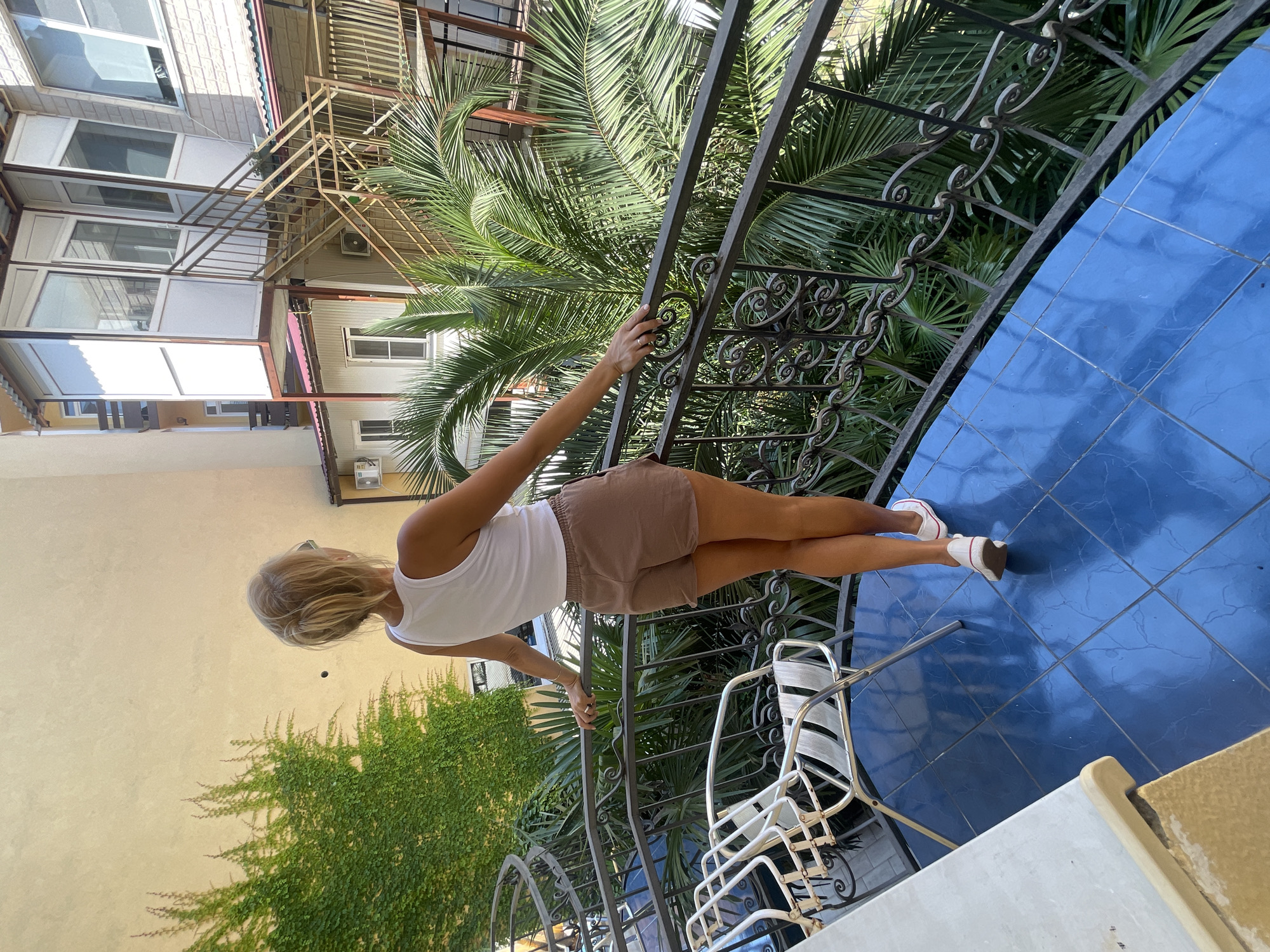 In Sochi, Russia, a woman with blonde hair tied in a bun, wearing a white tank top and brown shorts, stands on a balcony. She is facing away from the camera, looking out towards the surrounding environment. The balcony has a blue tiled floor and a dark metal railing with decorative scrollwork. Next to her on the balcony are two white metal chairs. Lush green palm fronds are visible behind the railing, suggesting a tropical or subtropical setting. In the background, there is a building with multiple levels and balconies, some enclosed with glass. The wall of an adjacent building is covered in vibrant green ivy. The scene captures a moment of relaxation or contemplation on a sunny day.