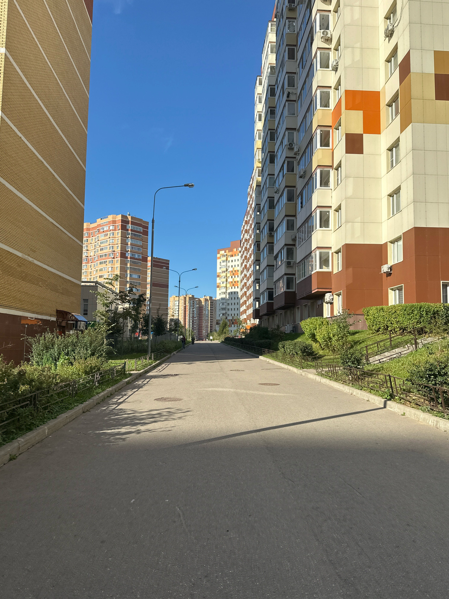 This is a full shot of a street in the city of Vidnoye, Russia, lined with multi-story residential buildings on both sides. The road is paved and empty except for a single person walking in the distance. The buildings are modern, with a mix of yellow, white, brown, and orange facades. Green trees and bushes are planted along the sides of the road, and there is a metal fence separating the sidewalk from the greenery. The sky is clear and blue, and the sun casts long shadows on the road. The overall impression is one of a well-maintained, urban residential area on a sunny day.