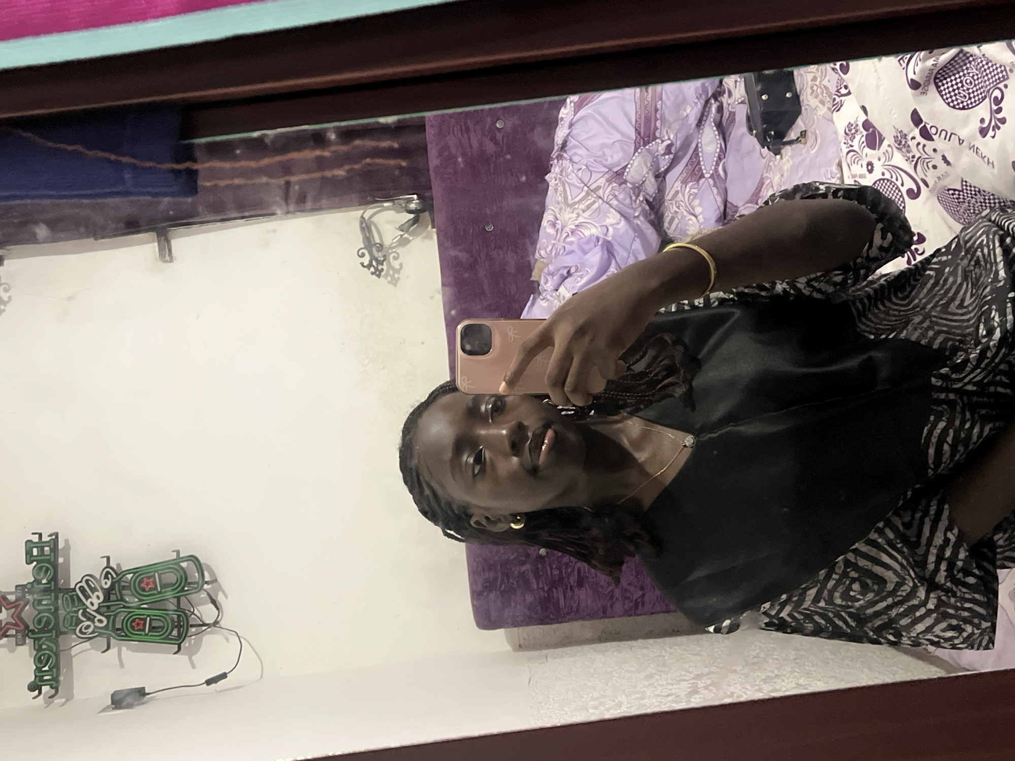 A young Black woman is taking a selfie in a mirror. She is holding a pink smartphone in her right hand. She is wearing a black top and a gold bracelet on her left wrist. She has braided hair and gold hoop earrings. Behind her is a bed with a purple headboard and patterned bedding. There is a Heineken neon sign on the wall to the left. The location appears to be a bedroom in an unknown location, possibly in Abuja, Nigeria.