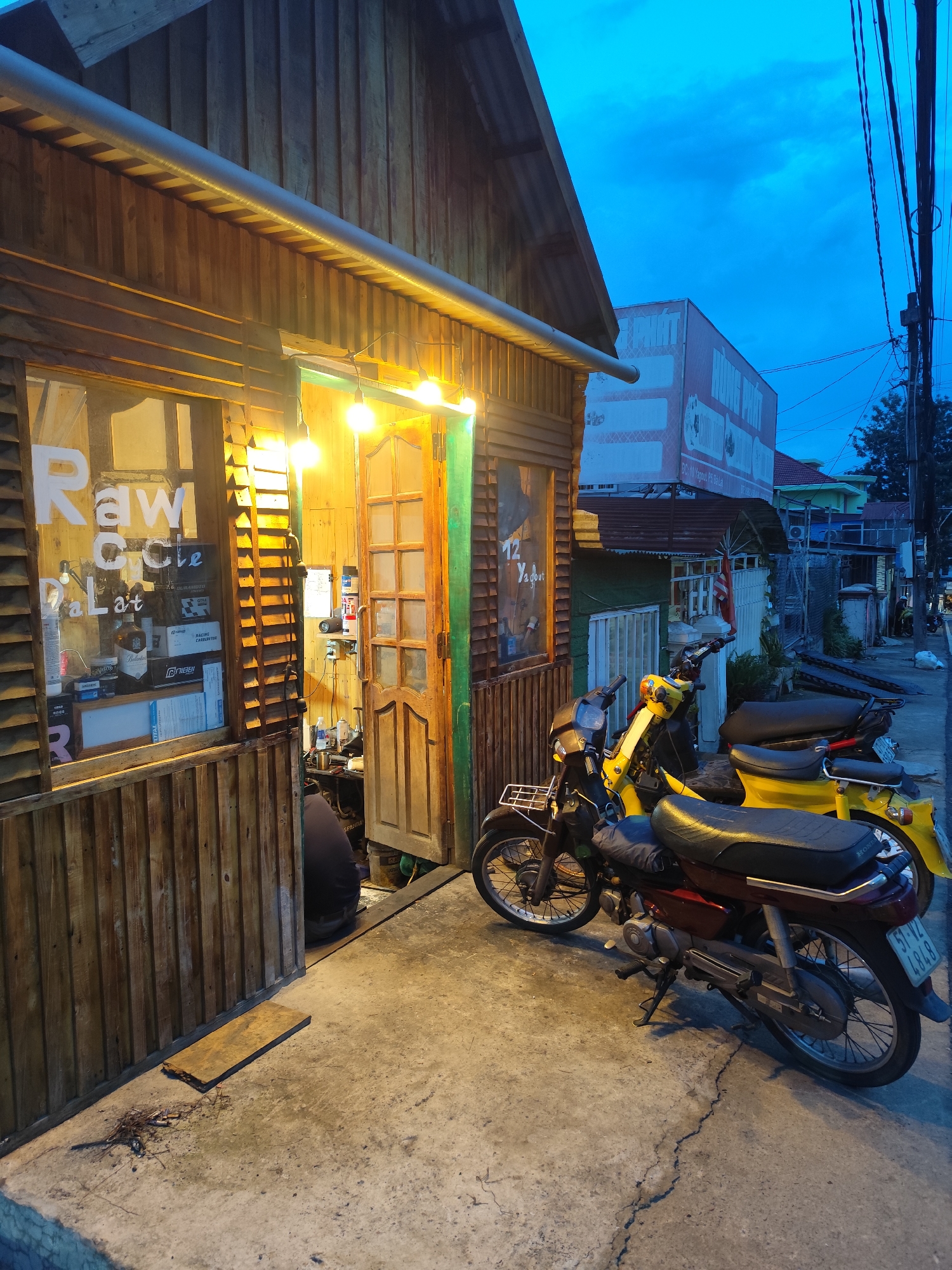 The image shows a wooden building with a sign that reads "Raw Coffee Dalat" on the window. There are several motorcycles parked in front of the building. The sky is dark blue, indicating that it is either dusk or dawn. The building appears to be a coffee shop or a similar establishment. There is a person inside the building, partially visible, appearing to be working on something. The street in front of the building is paved with concrete and has some cracks. The overall scene suggests a quiet, urban environment in Cam Ly - Đà Lạt, Vietnam.