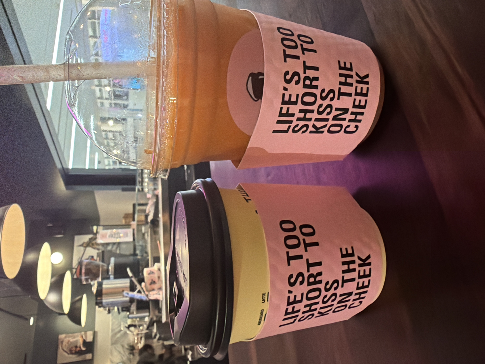 Two drinks, a coffee cup and a large plastic cup with a straw, sit on a dark, reflective surface. The coffee cup is yellow with a dark lid and a pink sleeve that reads "LIFE'S TOO SHORT TO KISS ON THE CHEEK". The plastic cup contains an orange beverage and also has a pink sleeve with the same text. The background appears to be an indoor establishment with hanging lights and a counter area. The context of Minneapolis, United States of America is not discernible from the image.